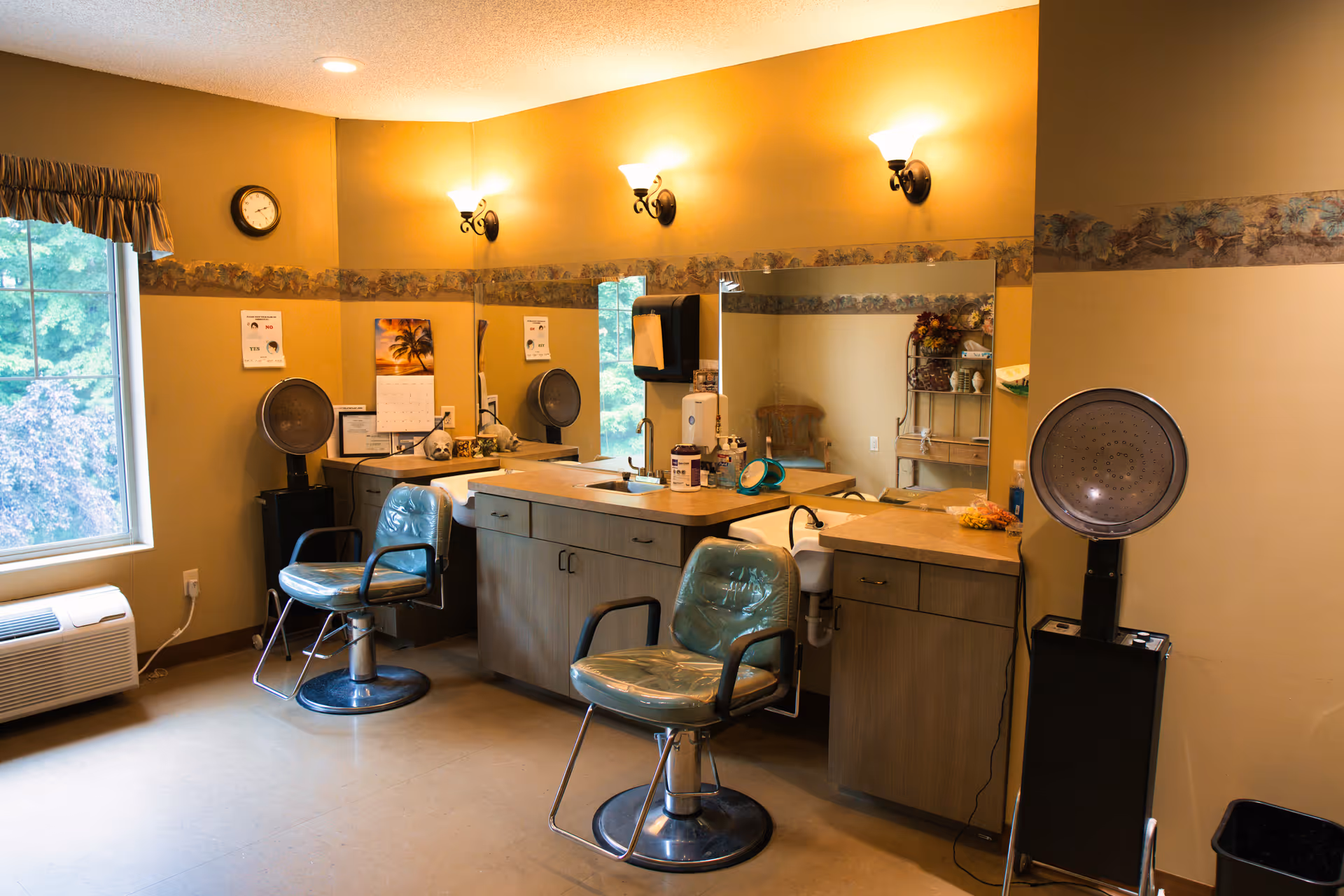 Small salon room with two styling chairs, hooded hair dryers, a large mirror and a countertop in a warm-toned interior.