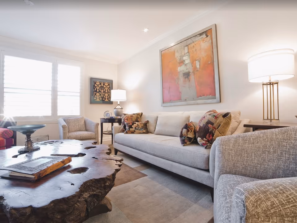 A bright and cozy living room featuring a light gray sofa with patterned throw pillows, two matching armchairs, a unique wooden coffee table with a natural edge, two side tables with lamps, and abstract artwork on the walls. Sunlight streams through white plantation shutters on the window.