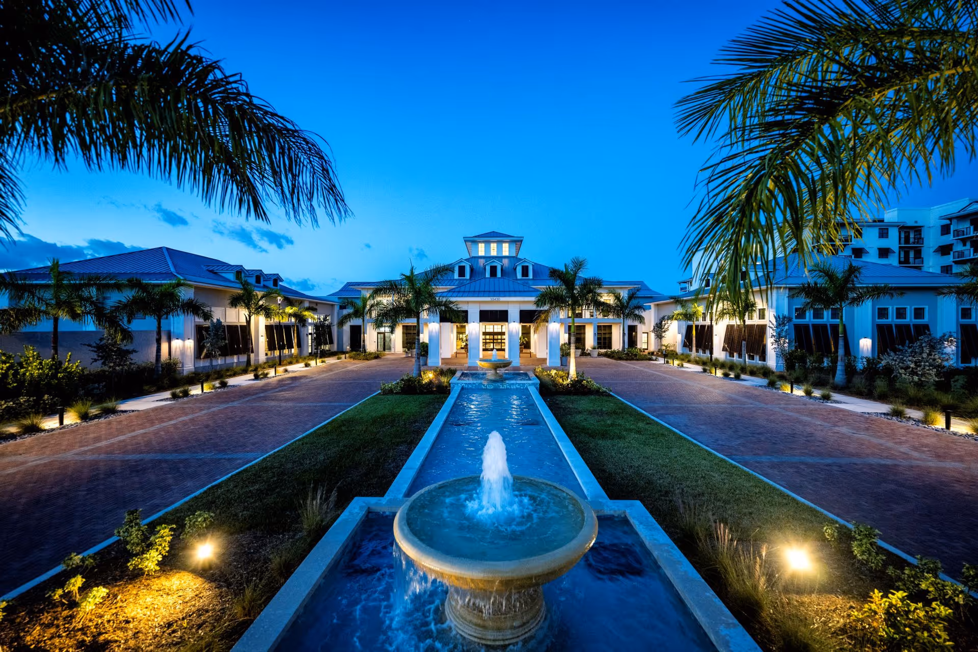 Front entrance of a senior living community at dusk with a central fountain, palm trees, and a lit driveway.