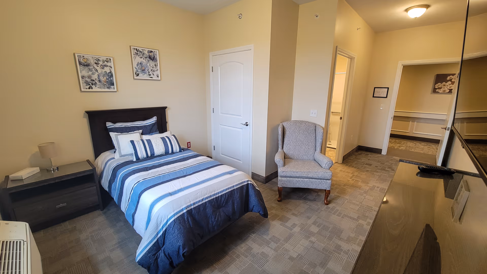 A neatly arranged bedroom with a single bed featuring blue and white striped bedding, a nightstand with a lamp and tissue box, a comfortable upholstered armchair, and a flat-screen TV mounted on the wall. The room has beige walls, carpeted flooring, and two floral paintings above the bed. There is a door leading to a bathroom and another open doorway leading to a hallway.