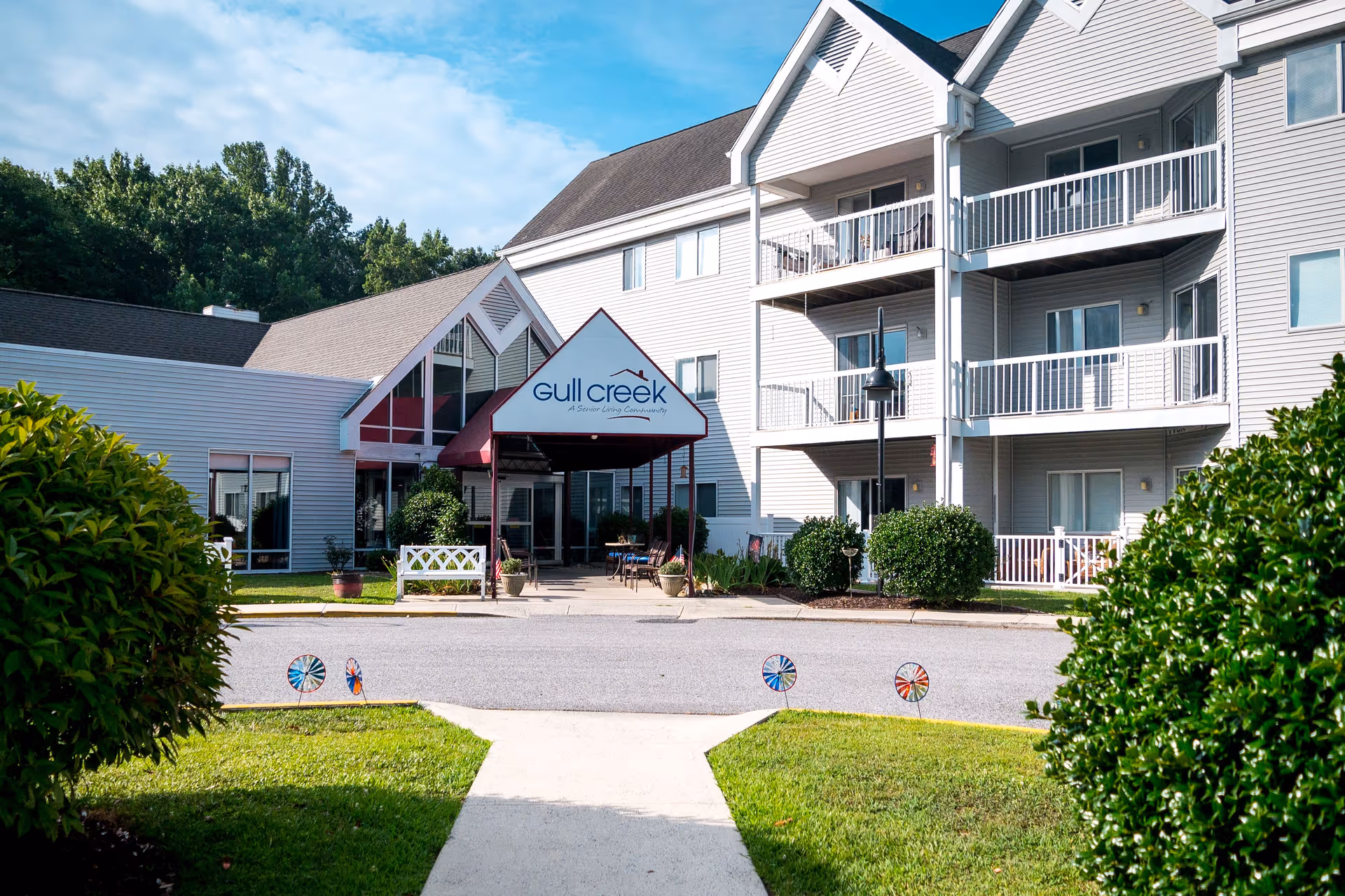 Exterior view of Gull Creek senior living community building with a covered entrance, balconies, green bushes, and a clear blue sky.