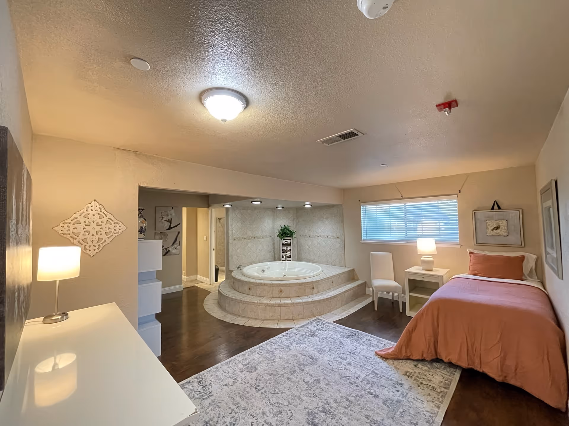 A cozy bedroom with a single bed covered in a coral blanket and a matching pillow. Next to the bed is a white nightstand with a lamp and a white chair. The room features a large built-in round jacuzzi tub with tiled steps leading up to it. The walls are beige, and there is a window with blinds letting in natural light. The floor is a combination of dark wood and a patterned area rug. Decorative wall art and a small plant near the tub add to the room's ambiance.