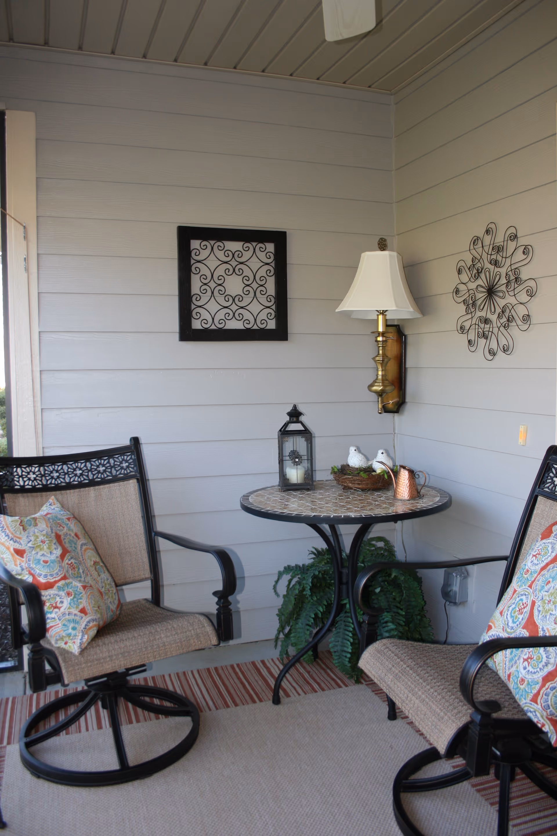 A covered patio seating area with two cushioned swivel chairs, a small round mosaic table with decorative items, a wall sconce and metal wall art.