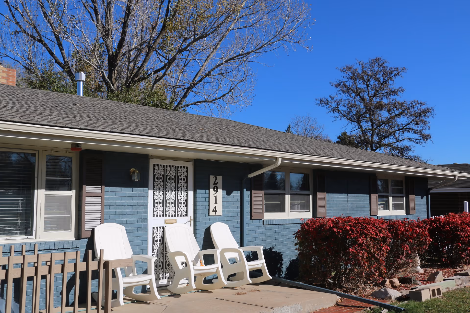 Single-story blue brick house front with white porch chairs, address number 2914, and red shrubs under a clear blue sky.