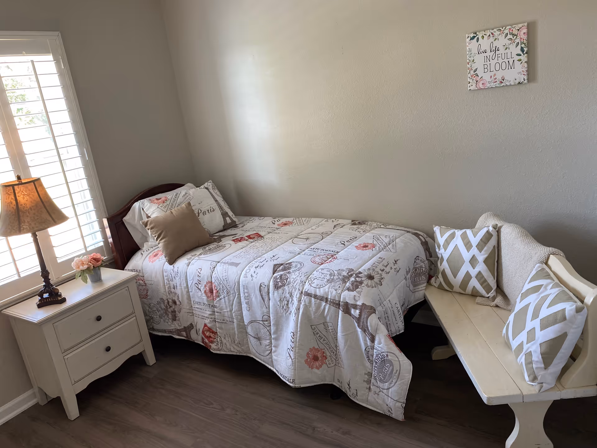 A small bedroom with a single bed covered in a Paris-themed quilt and pillows. Next to the bed is a white nightstand with two drawers, a decorative lamp, and a small vase with pink flowers. A window with white shutters is on the left wall. On the right side, there is a white wooden bench with two patterned pillows and a beige throw blanket. A wall decoration above the bench reads 'live life IN FULL BLOOM' with floral designs.