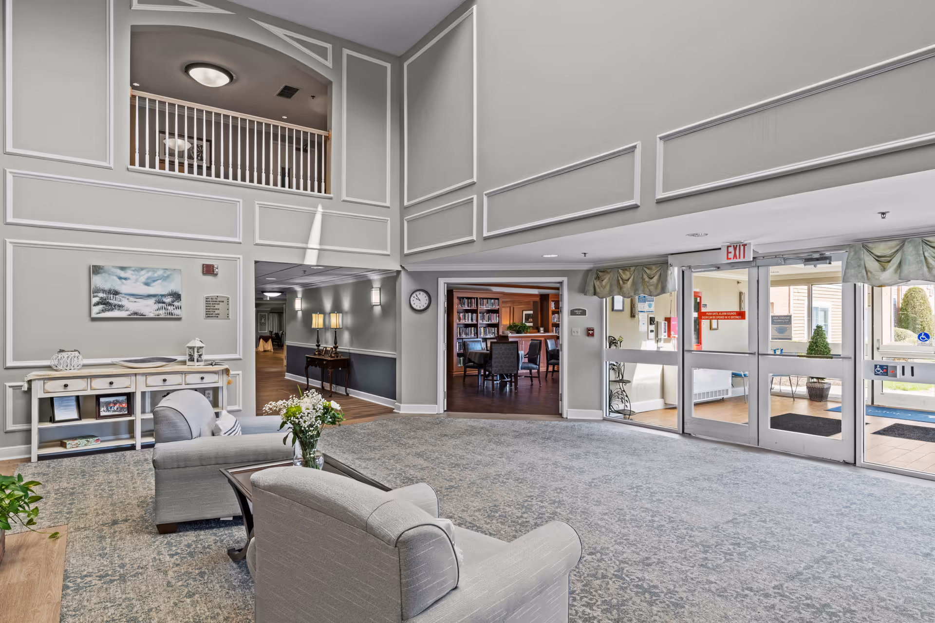 Spacious senior living facility lobby with high ceilings and decorative wall paneling. The area features two gray upholstered armchairs around a small table with a vase of flowers. To the left, there is a white console table with decorative items and a painting above it. A hallway with lamps on a side table leads to other rooms. Straight ahead, double doors open to a library or common room with bookshelves and chairs. On the right, glass exit doors lead outside, showing some greenery and outdoor seating.