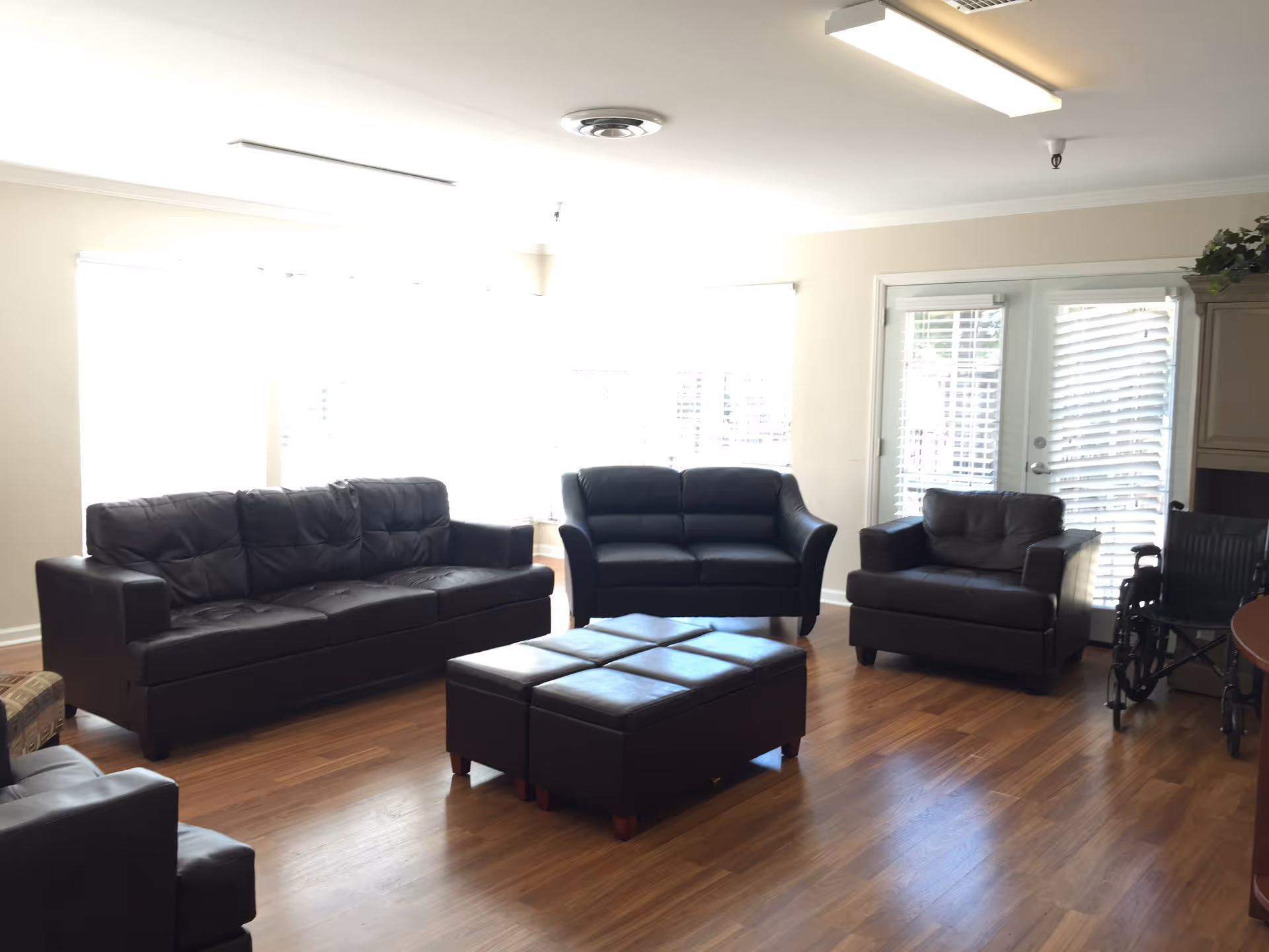 A bright living room with wooden flooring featuring three black leather sofas arranged around a central black leather ottoman. There is a wheelchair near a set of glass doors with white blinds, and a cabinet with a plant on top is visible in the corner.