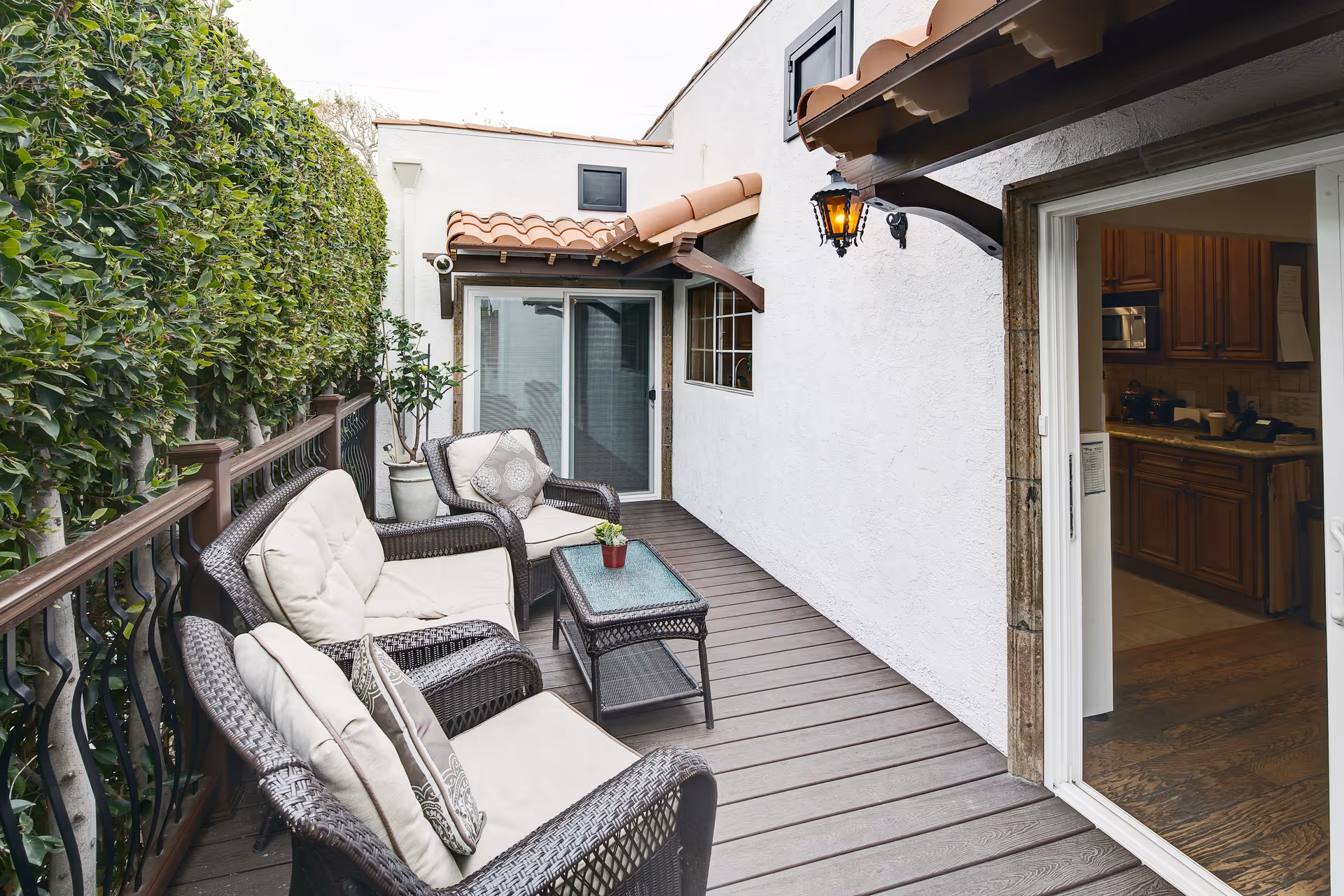Outdoor patio area with cushioned wicker chairs and a glass-top coffee table on a wooden deck. The patio is bordered by a tall hedge on one side and a white stucco wall with a lantern light fixture on the other. A sliding glass door leads to an interior kitchen with wooden cabinets.