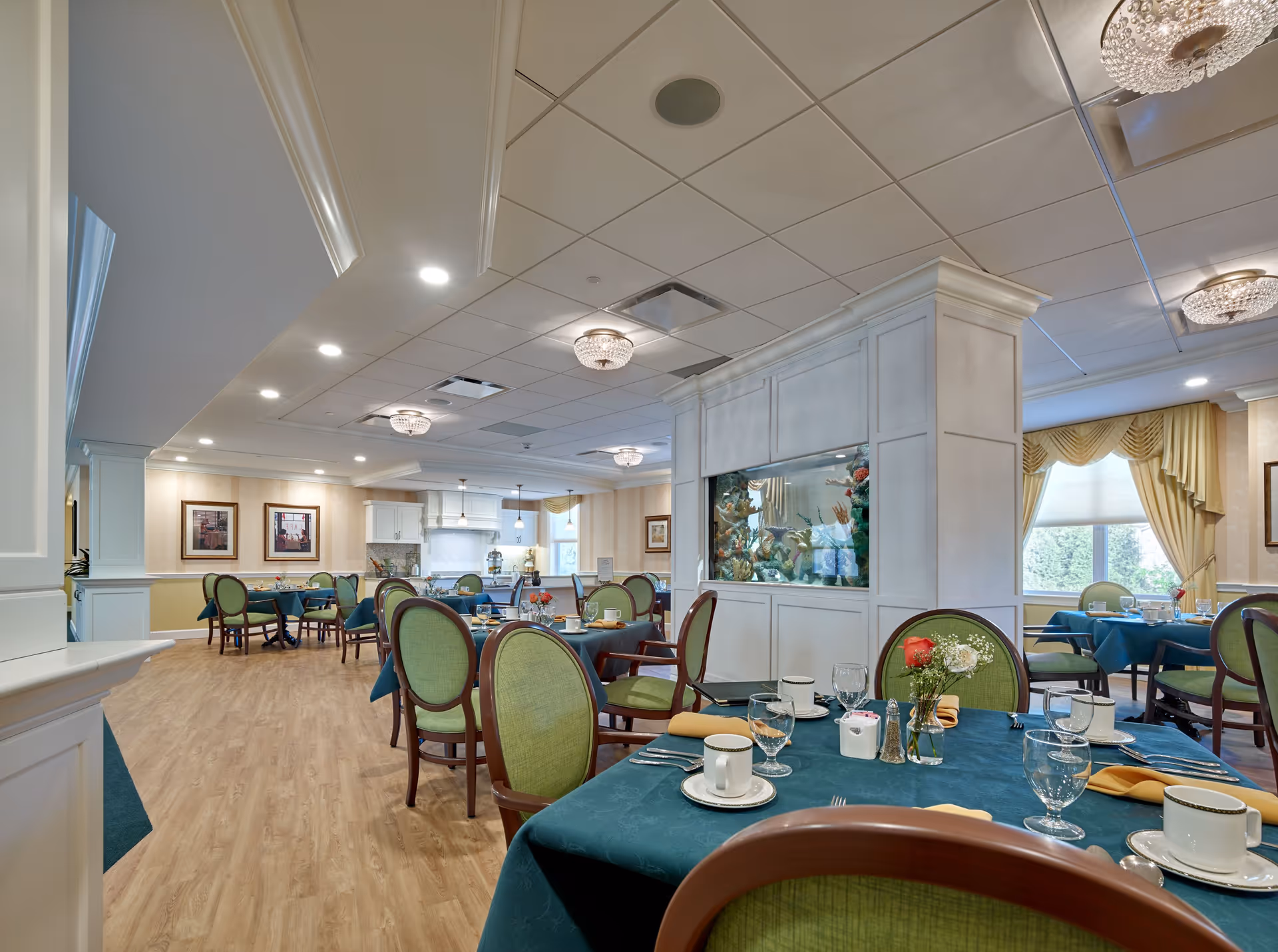 A dining room in a senior living facility with tables covered in teal tablecloths, set with cups, glasses, plates, and silverware. The chairs have green upholstery and wooden frames. There is a large built-in aquarium in the center of the room, and the room is well-lit with ceiling lights and natural light from windows with yellow curtains.