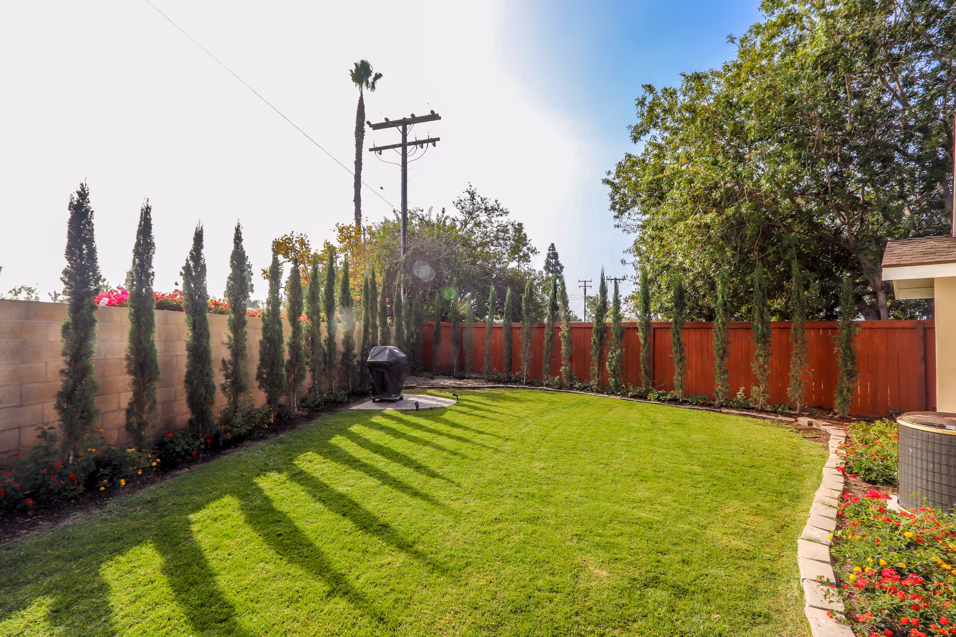 A well-maintained outdoor garden area with a green lawn, tall narrow trees lined along a brick wall and a wooden fence, a barbecue grill on a small concrete pad, and colorful flowers bordering the lawn under a clear blue sky.