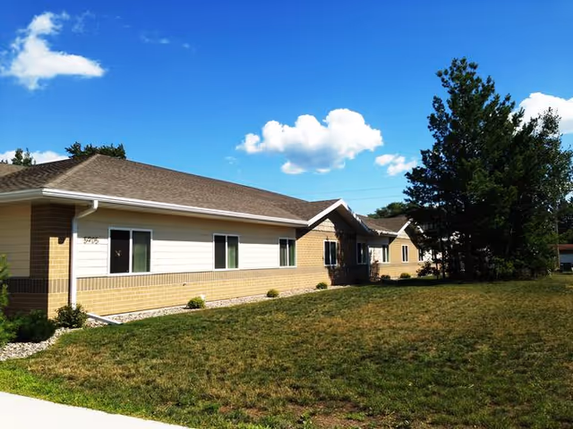 Exterior view of a single-story senior living facility building with beige brick and light siding, several windows, a sloped roof, and a grassy lawn with trees under a blue sky with scattered clouds.