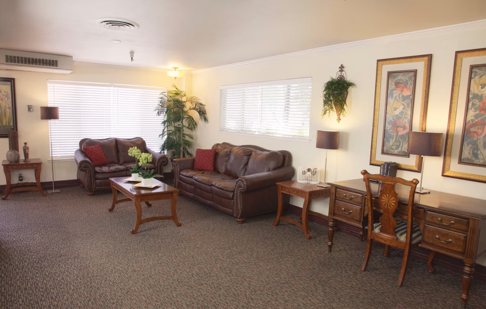 A cozy living room area with two brown leather sofas, a wooden coffee table with a plant centerpiece, a wooden desk with a chair, two table lamps, framed floral artwork on the walls, a potted plant in the corner, and windows with white blinds letting in natural light.