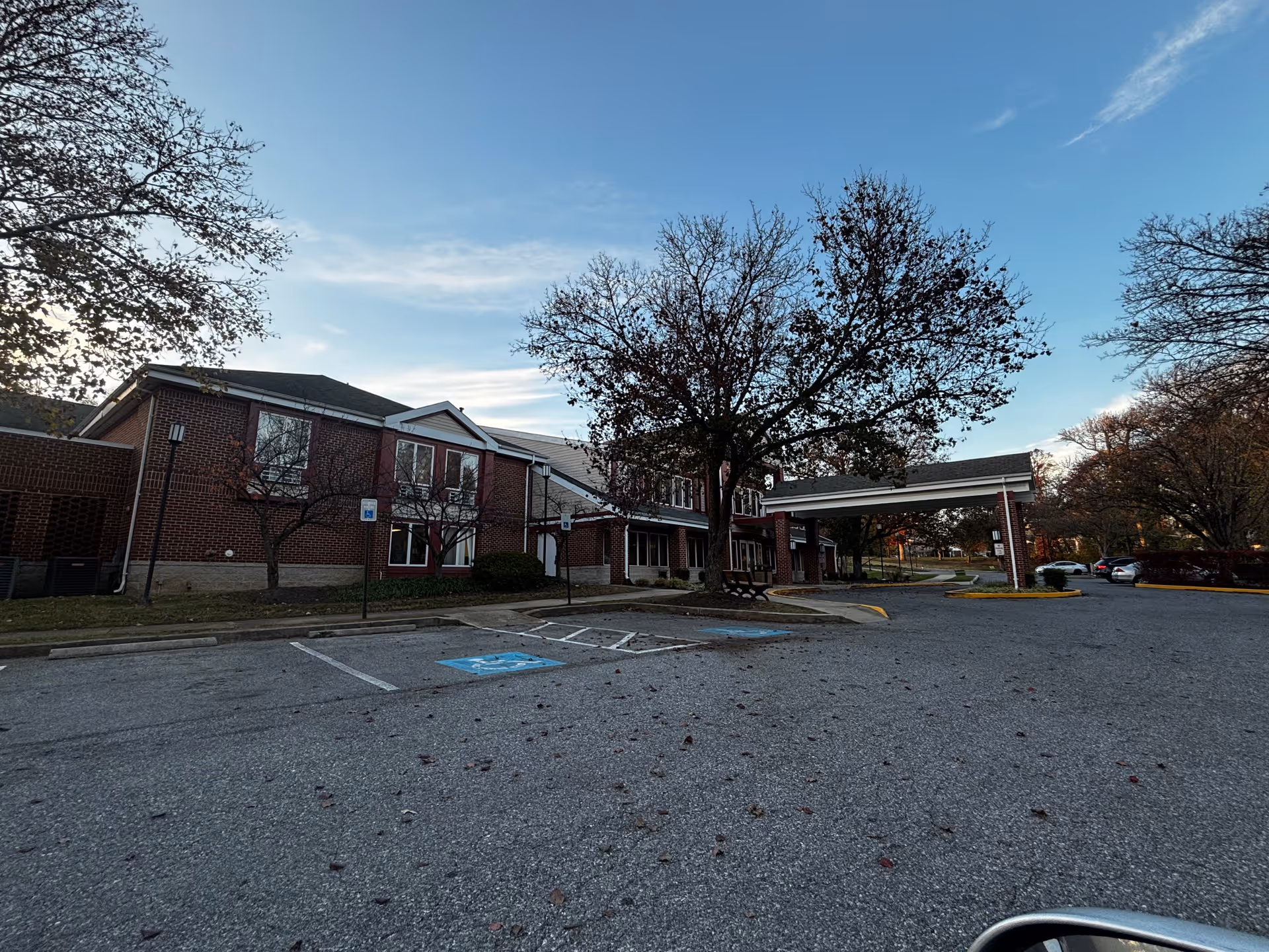 Exterior view of a two-story brick building with a covered entrance and several windows, surrounded by leafless trees and a parking lot with handicap parking spaces under a clear blue sky.
