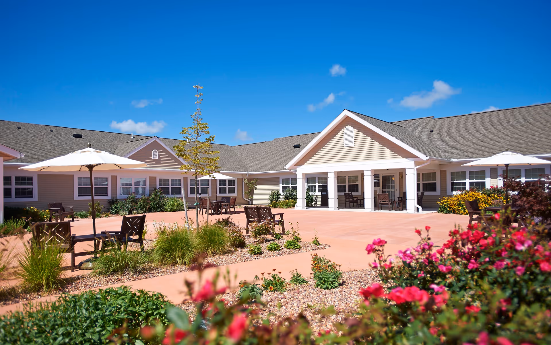 Outdoor courtyard area of a senior living facility with patio tables, chairs, and umbrellas surrounded by landscaped plants and flowers under a clear blue sky.