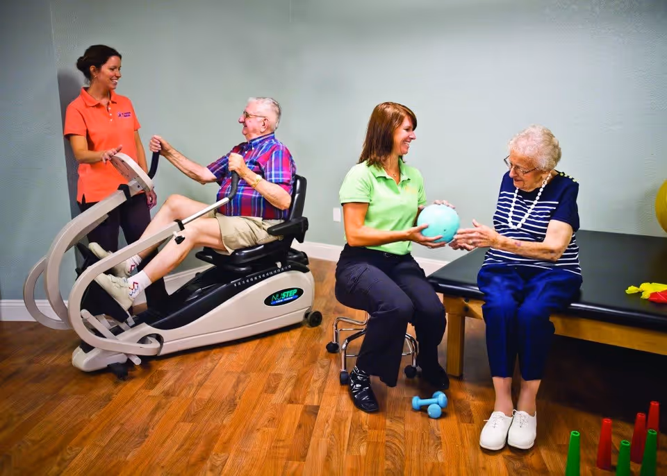 An elderly man exercises on a NuStep machine with the assistance of a female staff member in an orange shirt. Nearby, another female staff member in a green shirt is handing a light blue exercise ball to an elderly woman sitting on a padded table. The room has wooden flooring and light-colored walls.