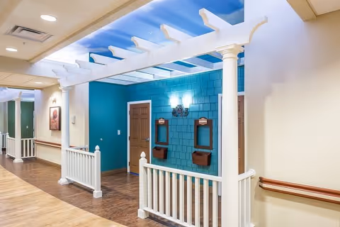 Interior hallway of a senior living facility with a white pergola structure, teal accent wall, two wooden doors, two wall-mounted mirrors with small shelves beneath, and wood flooring. The ceiling has recessed lighting and the walls are painted beige with wooden handrails.