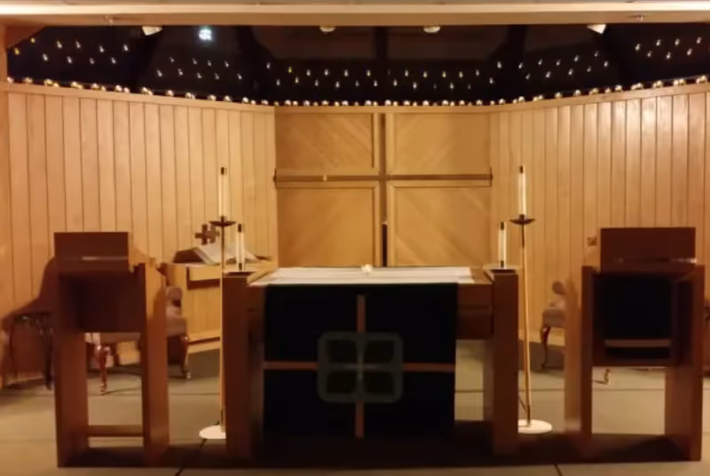 Interior view of a chapel or worship area with a wooden altar at the center, two wooden lecterns on either side, tall candles, and a large wooden cross mounted on the wood-paneled wall behind the altar.