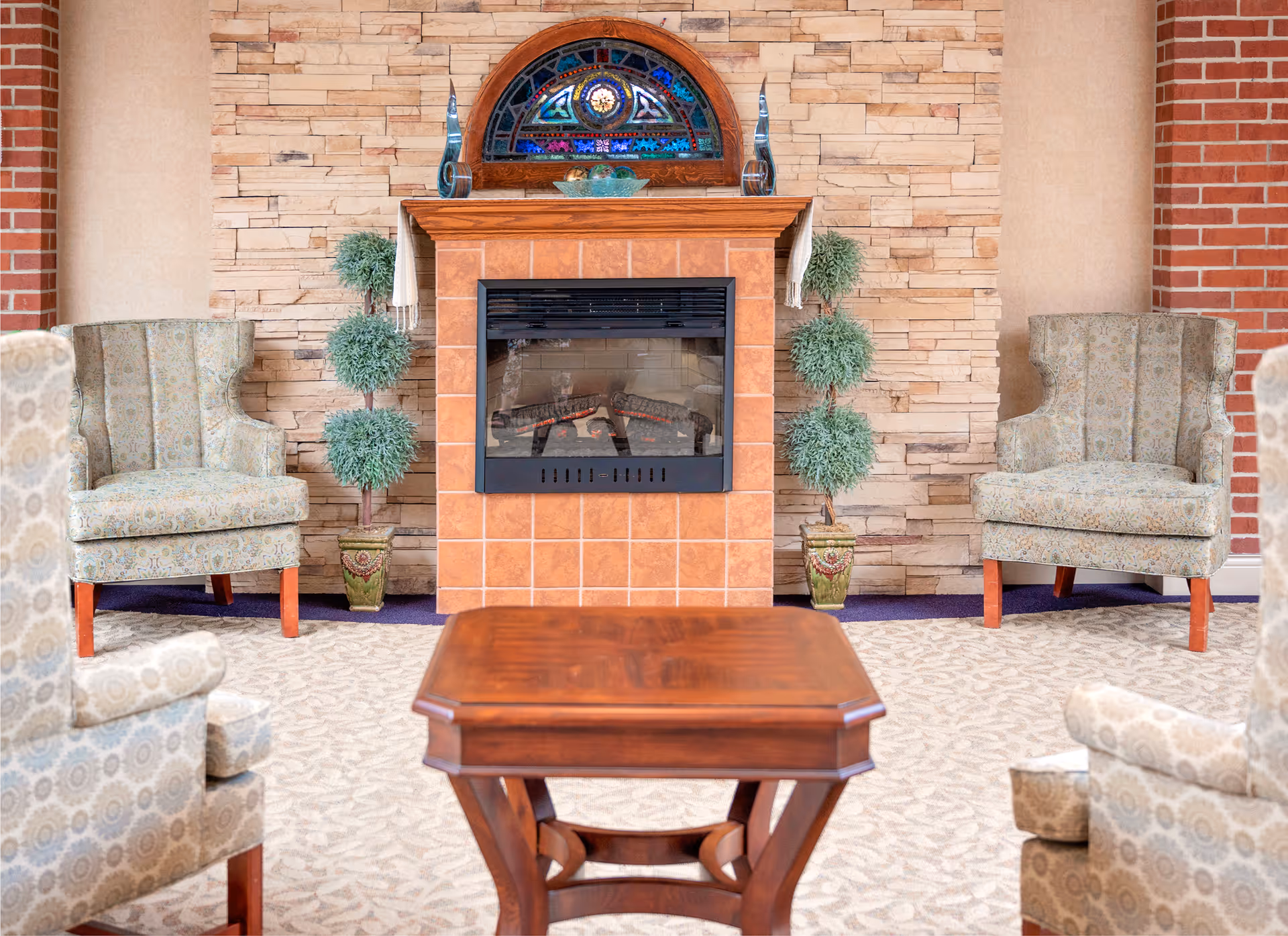 A cozy seating area with four patterned armchairs arranged around a wooden coffee table. In the background, there is a tiled fireplace with a decorative stained glass piece above it and two potted topiary plants on either side. The walls are a combination of brick and stone.