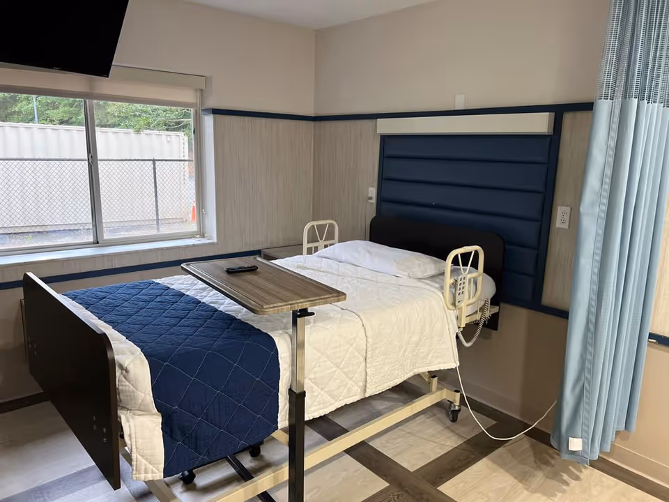 A hospital-style bed with white and blue bedding in a room with a large window, a mounted TV, a blue padded headboard, and a light blue privacy curtain partially drawn to the side. There is a small overbed table with a remote control on it.