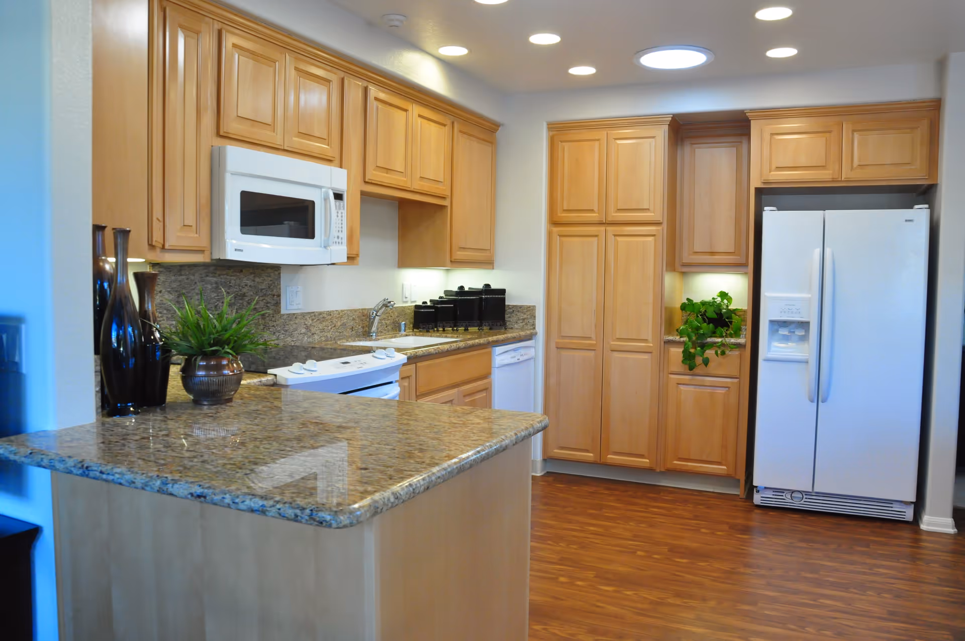 A modern kitchen with wooden cabinets, granite countertops, a white microwave, stove, dishwasher, and refrigerator. There are decorative vases and a potted plant on the counter, with wooden flooring and bright lighting.