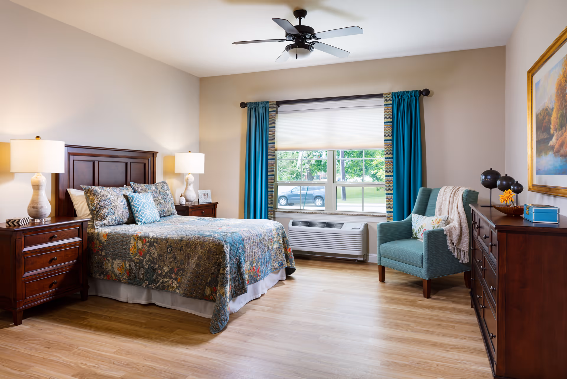 A well-lit bedroom with a wooden bed frame, floral patterned bedding, two bedside tables with lamps, a teal armchair with a throw blanket and pillow, a wooden dresser with decorative items, a large window with teal curtains, and a ceiling fan.