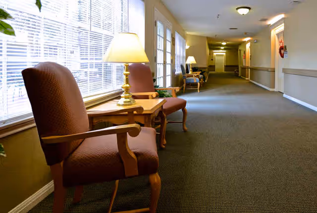 A long hallway in a senior living facility with carpeted floor, beige walls, and several wooden armchairs with cushions placed along the wall. Small tables with lamps are positioned between the chairs. Large windows on the left side allow natural light to fill the space.