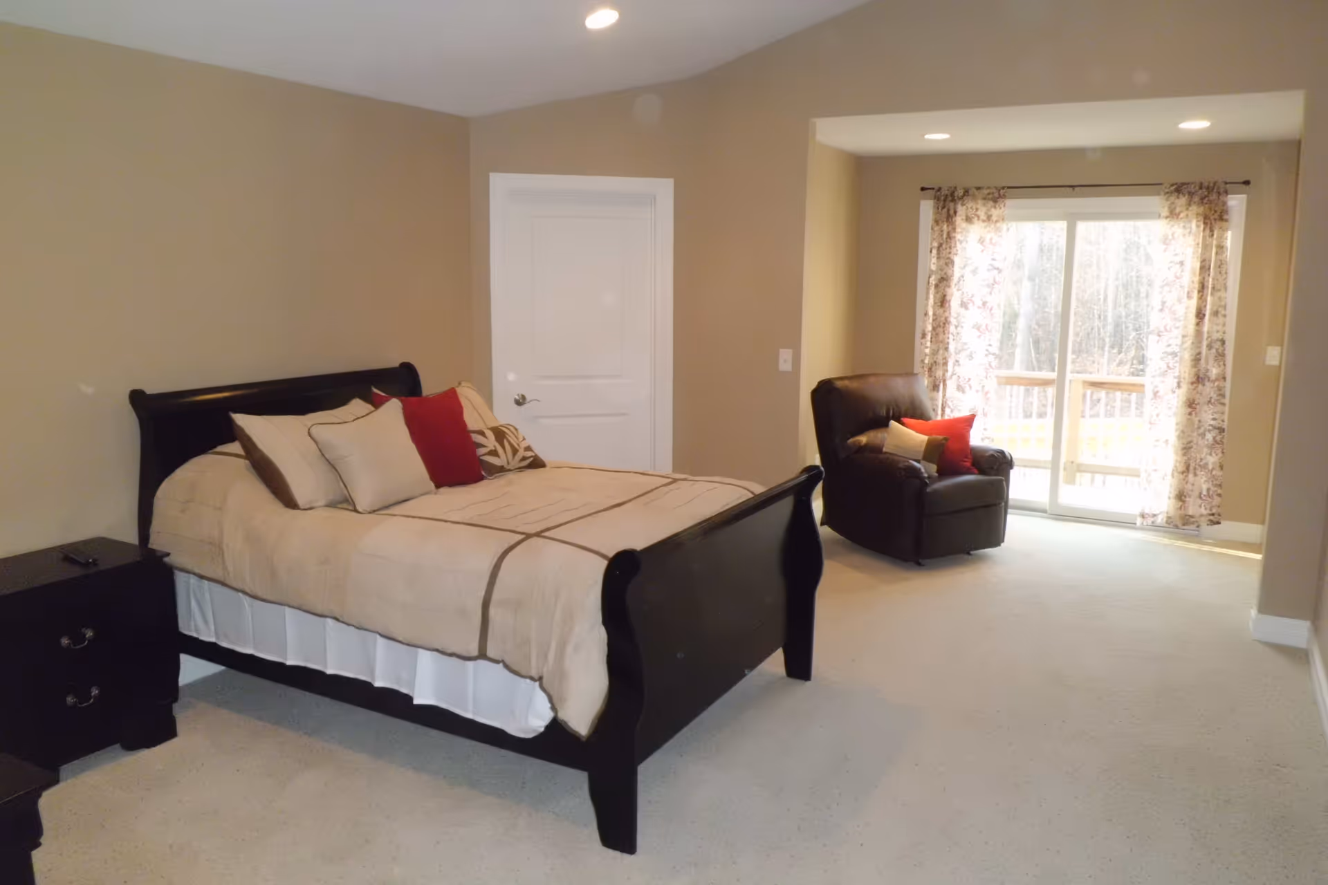 A bedroom with a dark wooden bed frame and beige bedding accented with multiple pillows in beige, brown, and red. There is a dark wooden nightstand with two drawers next to the bed. In the corner near a sliding glass door with floral curtains, there is a dark brown armchair with red and beige pillows. The room has beige walls and carpeted floor.