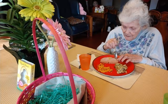 An elderly woman sitting at a dining table eating a meal consisting of sliced meat, corn, and a vegetable side. The table has a yellow tablecloth, a pink basket with green shredded paper, a vase with yellow and pink flowers, and a box of Russell Stover chocolates. In the background, there are chairs and other furniture in a common area.