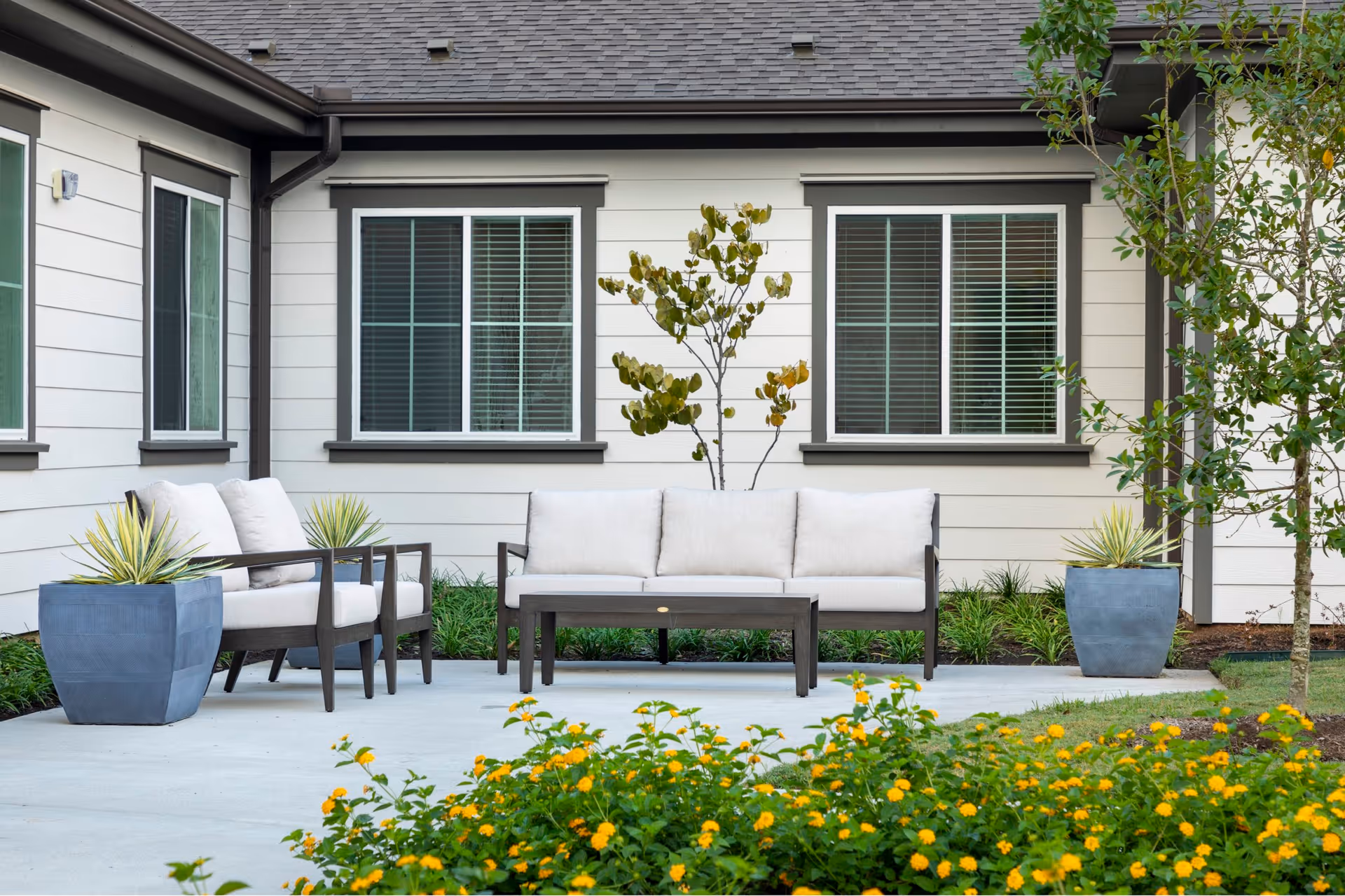 Outdoor patio area at The Landing at Augusta Woods Senior Living featuring cushioned seating including a sofa and two armchairs, a small coffee table, potted plants, and yellow flowers in the foreground with the building's exterior and windows in the background.
