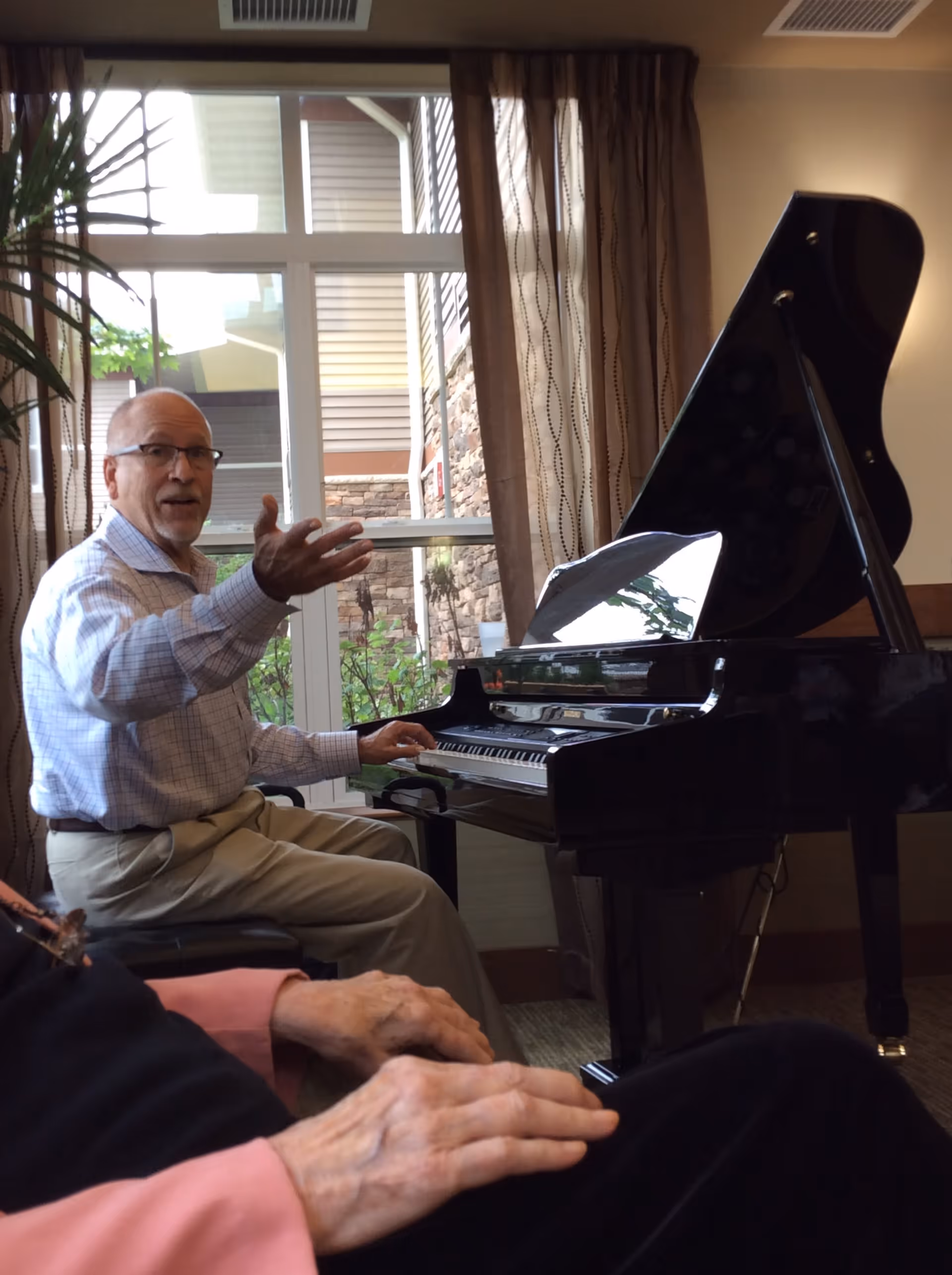 An elderly man wearing glasses and a checkered shirt is seated at a grand piano, gesturing with his right hand as if speaking or playing music. In the foreground, the hands of another elderly person are visible resting on their lap. The room has large windows with sheer curtains, allowing natural light to illuminate the space.