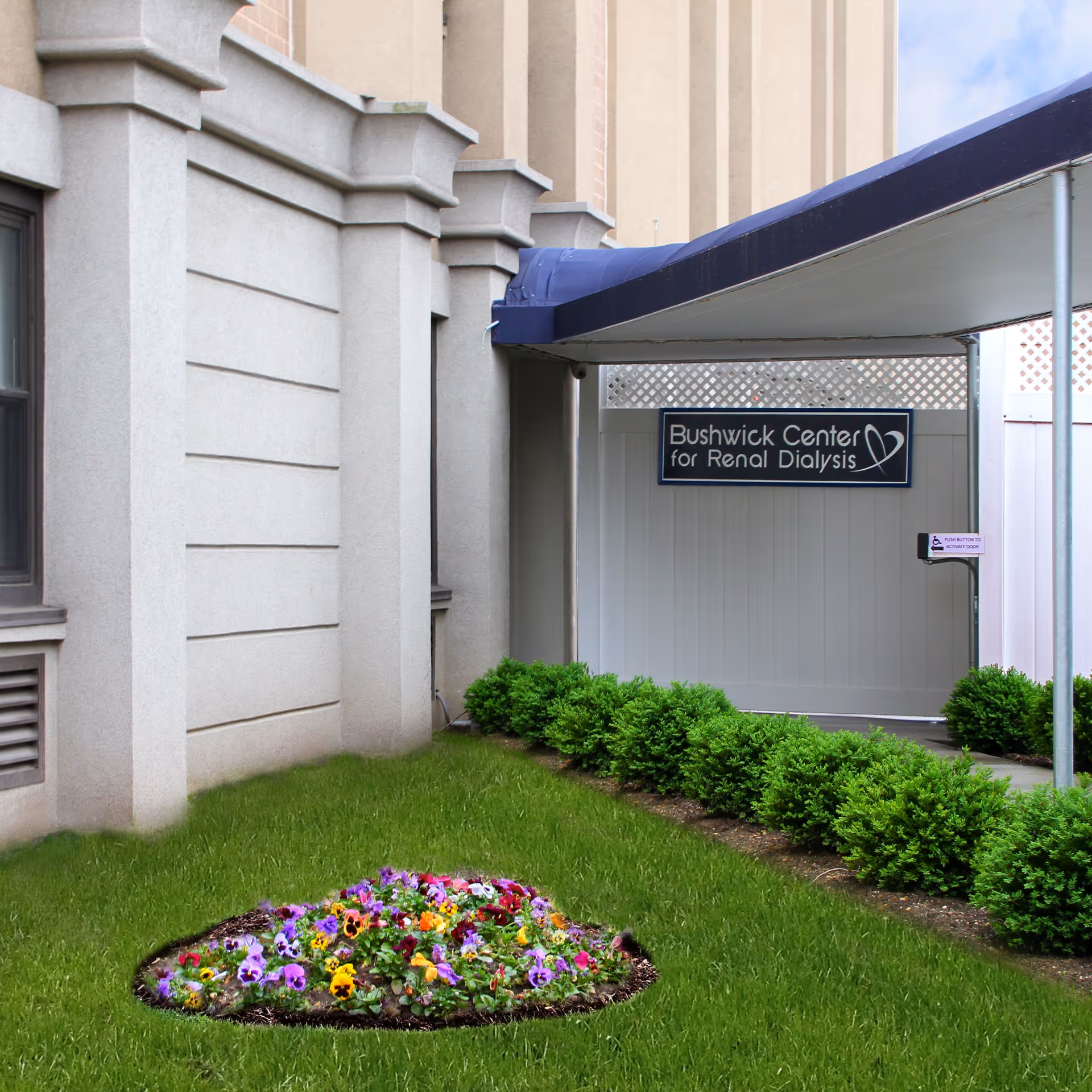 Covered entrance to the Bushwick Center for Renal Dialysis at a building front with a lawn, shrubbery, and a flowerbed.