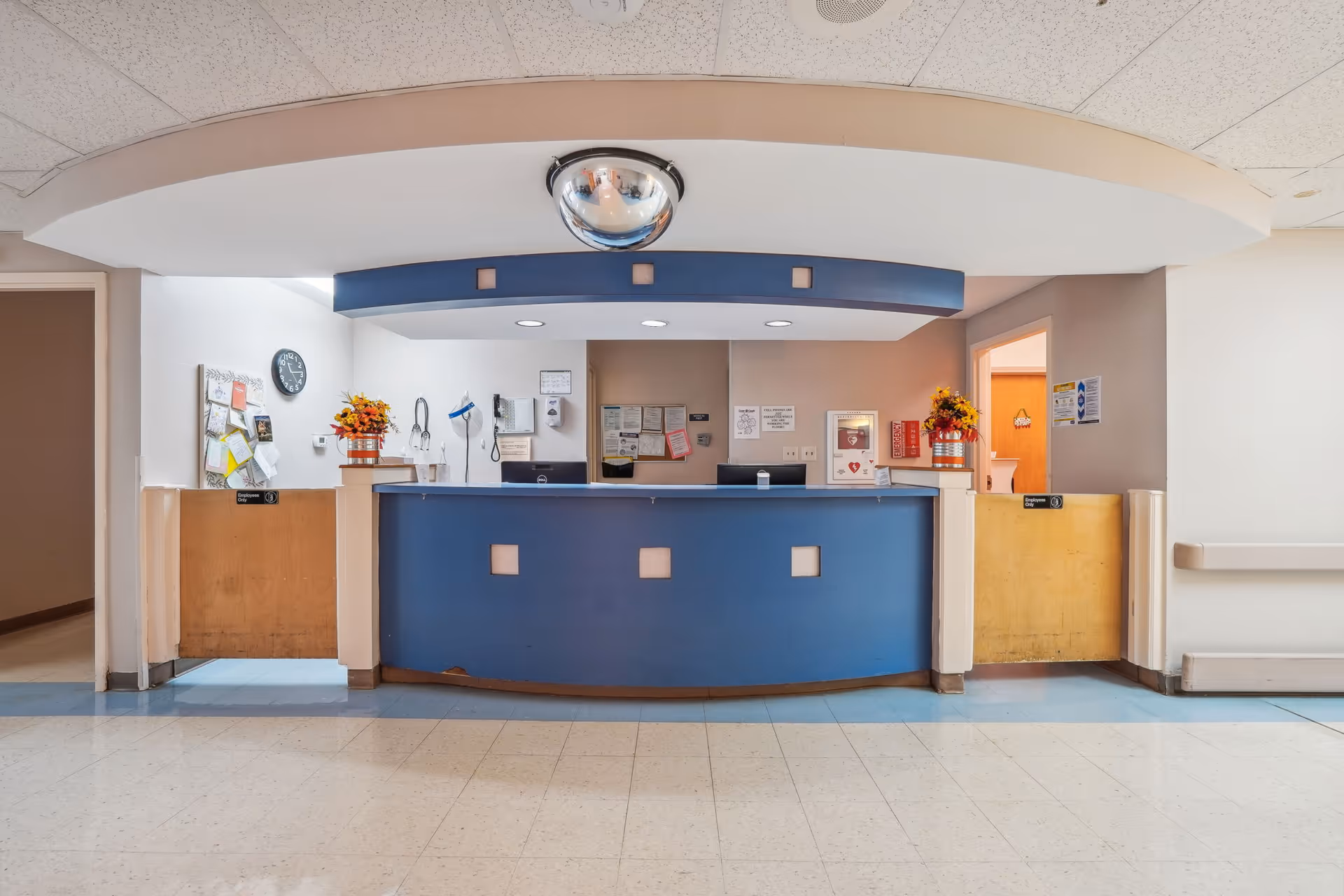 Reception desk area in a healthcare facility with a blue counter, bulletin boards with notices, a clock on the wall, and flower arrangements on either side. The space has a tiled floor and a curved ceiling feature with a convex security mirror above the desk.