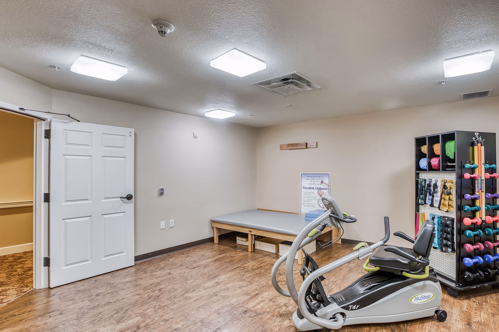 A small fitness and therapy room with a NuStep recumbent exercise machine, a rack of colorful dumbbells and weights, and a padded therapy table.