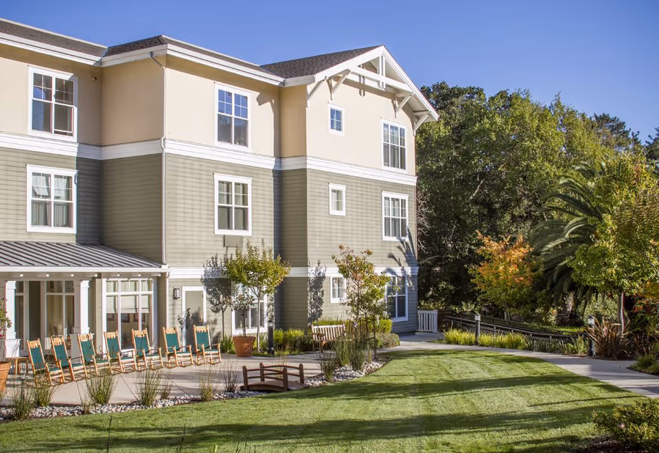 Three-story senior living building with a patio of rocking chairs, manicured lawn, and landscaped trees under a clear blue sky.