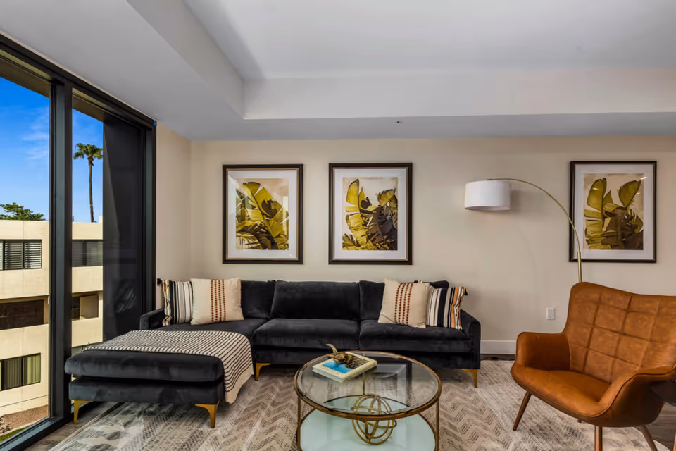 Modern living room with a black sectional sofa adorned with striped and patterned pillows, a round glass coffee table with decorative items, a brown leather armchair, three framed botanical prints on the wall, a floor lamp with a white shade, and large windows showing an exterior view with palm trees and a neighboring building.