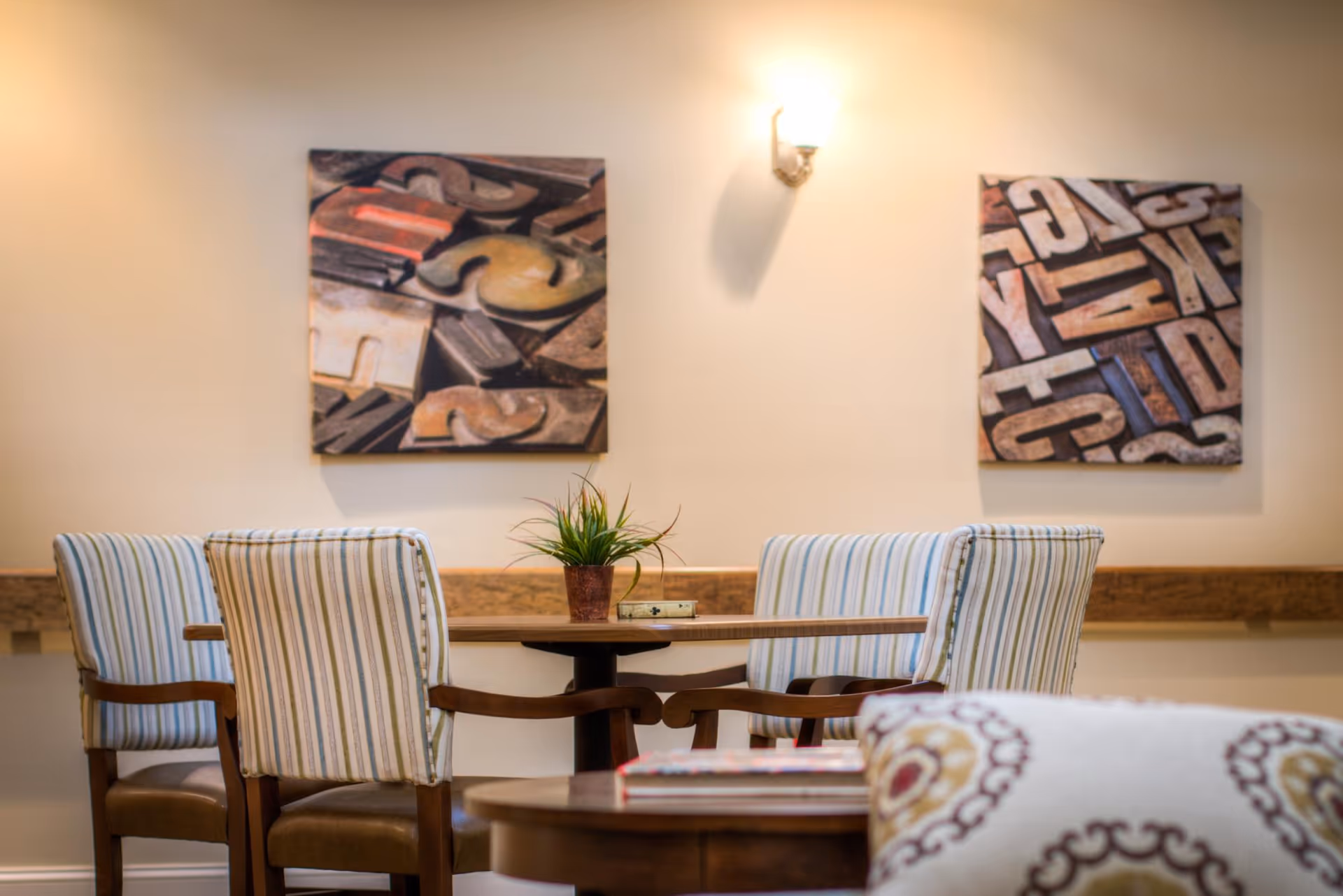 A cozy seating area with a wooden table surrounded by four striped upholstered chairs. On the table is a small potted plant and a book. The wall behind features two framed abstract artworks with letterpress blocks, and a wall sconce light fixture above.