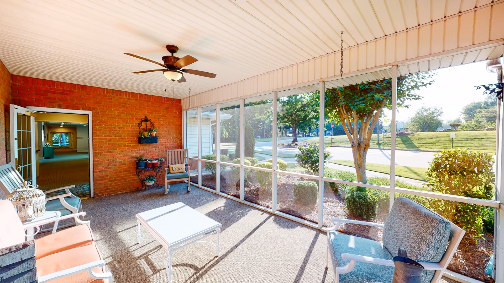 A bright screened-in porch area with comfortable seating including chairs and a small table. The porch has a ceiling fan and overlooks a landscaped outdoor area with trees and bushes. A door leads inside to a hallway with carpeted floors.