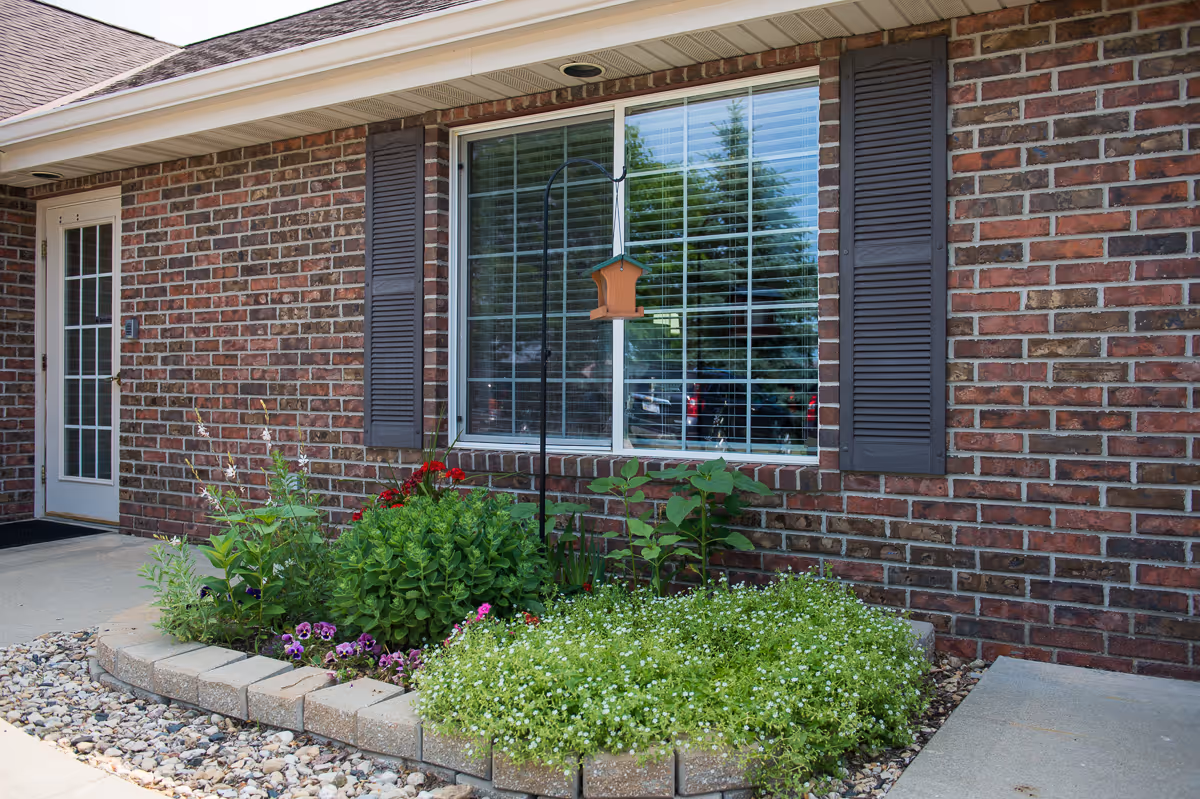 Exterior view of a brick building with a large window featuring closed blinds and dark shutters on either side. Below the window is a small garden bed bordered with bricks, containing various green plants and flowers. A bird feeder hangs on a metal pole in front of the window. To the left is a white door with glass panes.