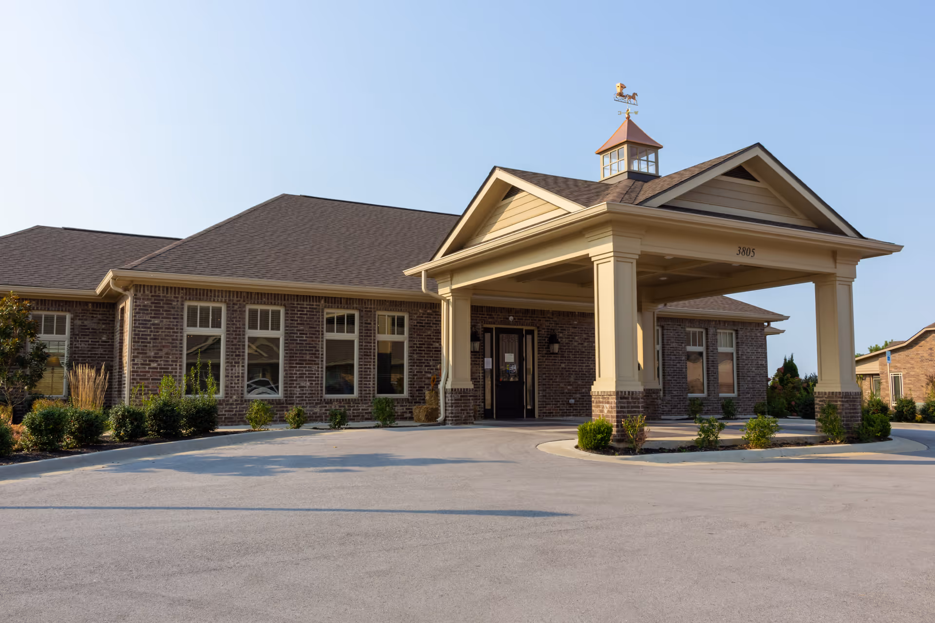 Exterior view of a single-story brick building with a covered entrance supported by columns. The building has multiple windows and a small cupola with a weather vane on the roof. There are small bushes and landscaping around the building and a clear blue sky above.