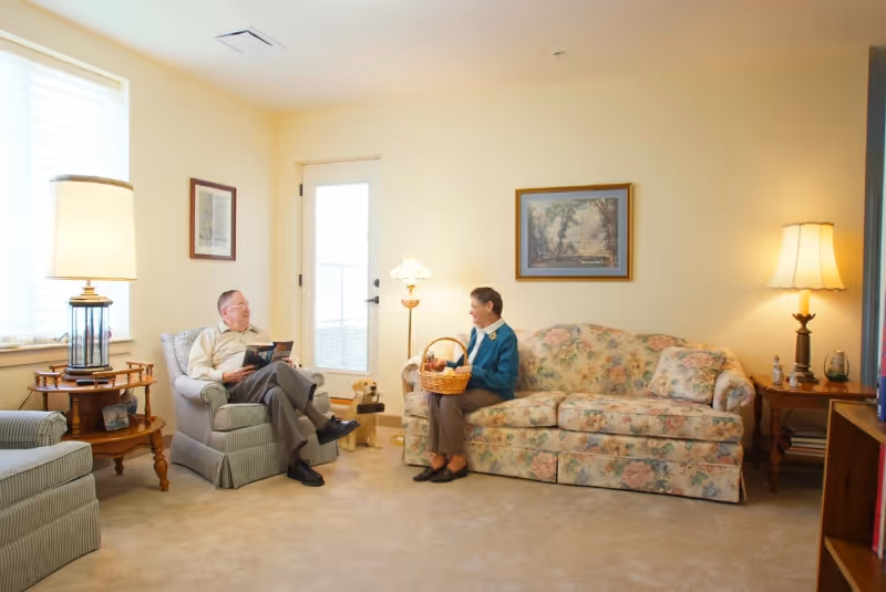A cozy living room with two elderly people sitting and conversing. One person is seated in a striped armchair reading a book, while the other sits on a floral-patterned sofa holding a basket. The room is warmly lit with two lamps, has framed artwork on the walls, and a door leading outside.