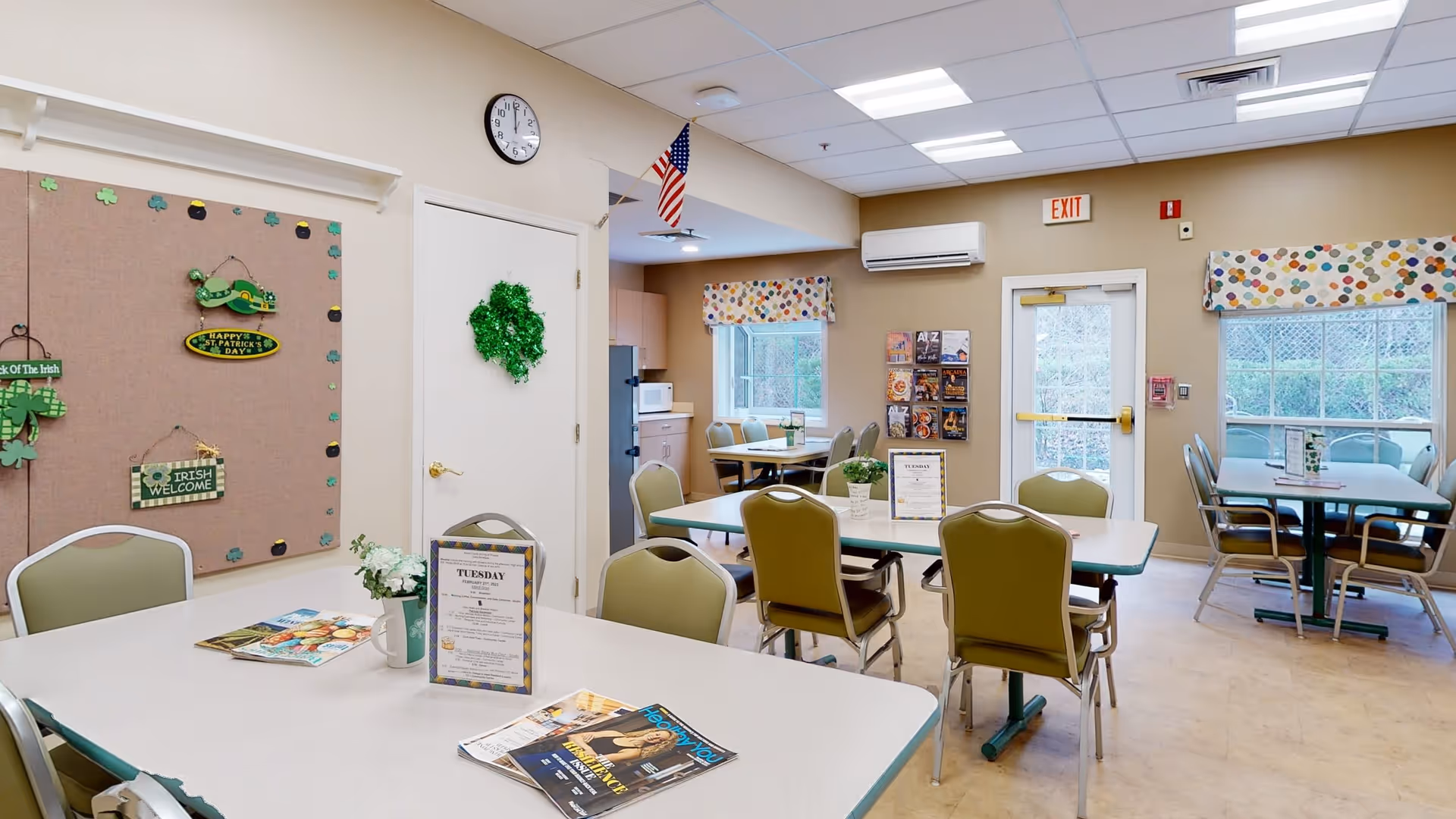 A bright dining room in a senior living facility with several tables and chairs arranged neatly. The walls are decorated with St. Patrick's Day themed decorations, including shamrocks and signs. There are windows with colorful valances, a clock on the wall, and an American flag hanging near the ceiling. A bulletin board and a rack with brochures are visible near the exit door.