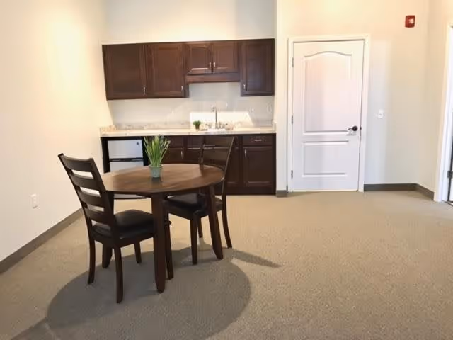 A small kitchenette area with dark wooden cabinets, a countertop with a sink, and a mini refrigerator. In front of the kitchenette is a round wooden table with two matching chairs and a small potted plant on the table. The room has beige carpet and light-colored walls with a white door on the right side.