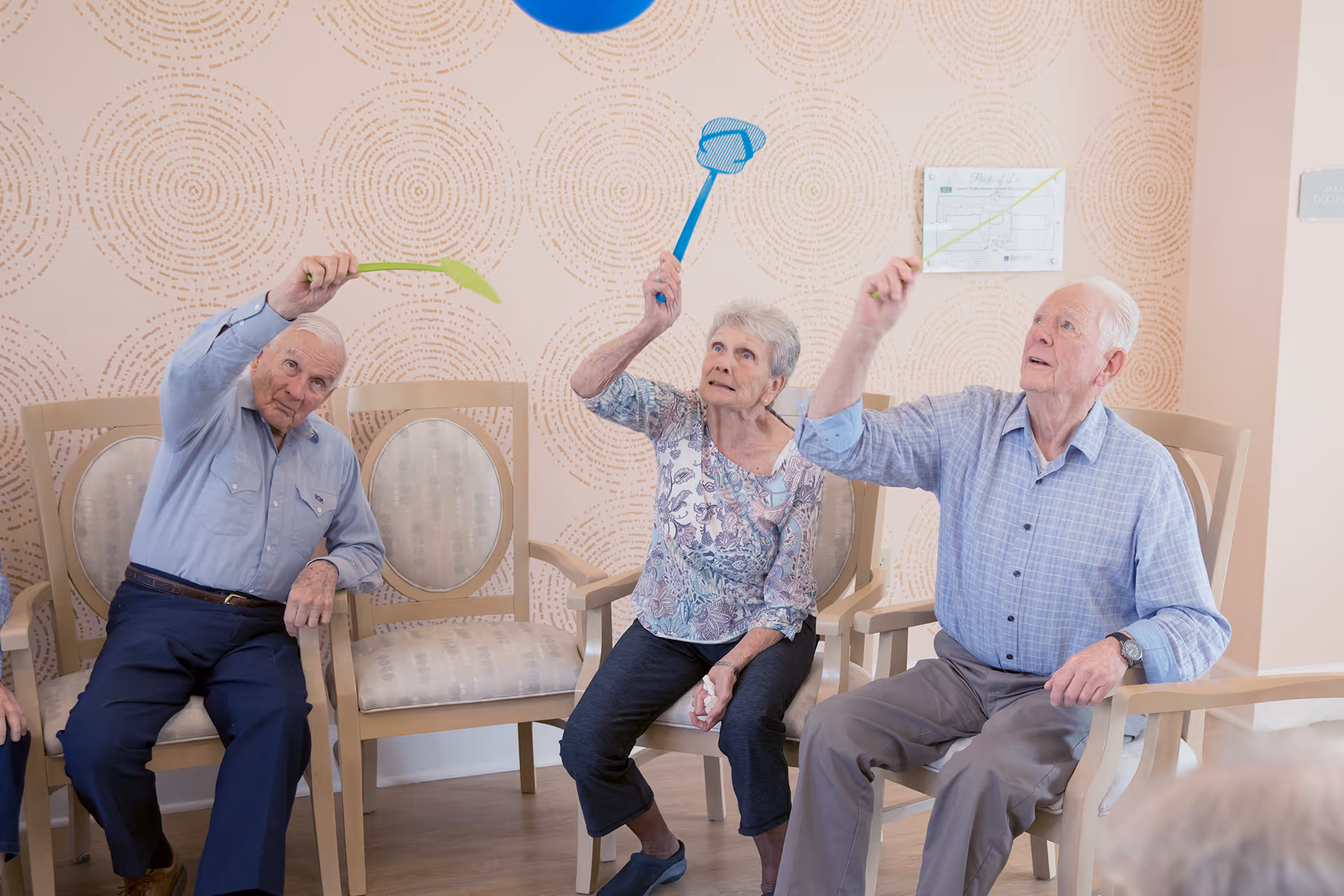 Three elderly individuals sitting on chairs in a room with patterned wallpaper, each holding a colorful stick and reaching upwards towards a blue balloon above them.