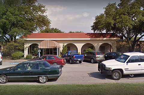 Exterior view of a single-story building with a red-tiled roof and arched entryways, several cars parked in front, and large trees on either side.