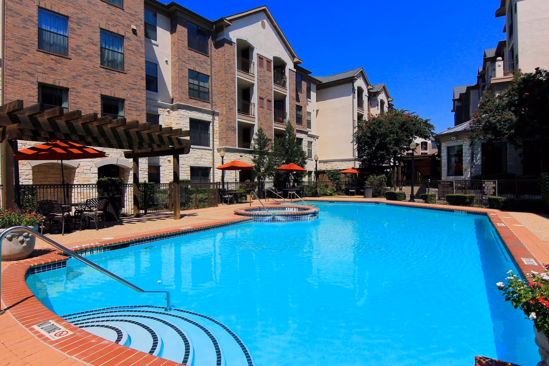 Outdoor swimming pool area at Conservatory At North Austin with clear blue water, surrounded by brick and stone buildings. There are several red umbrellas providing shade over seating areas, a wooden pergola, and some greenery including trees and potted plants under a bright blue sky.