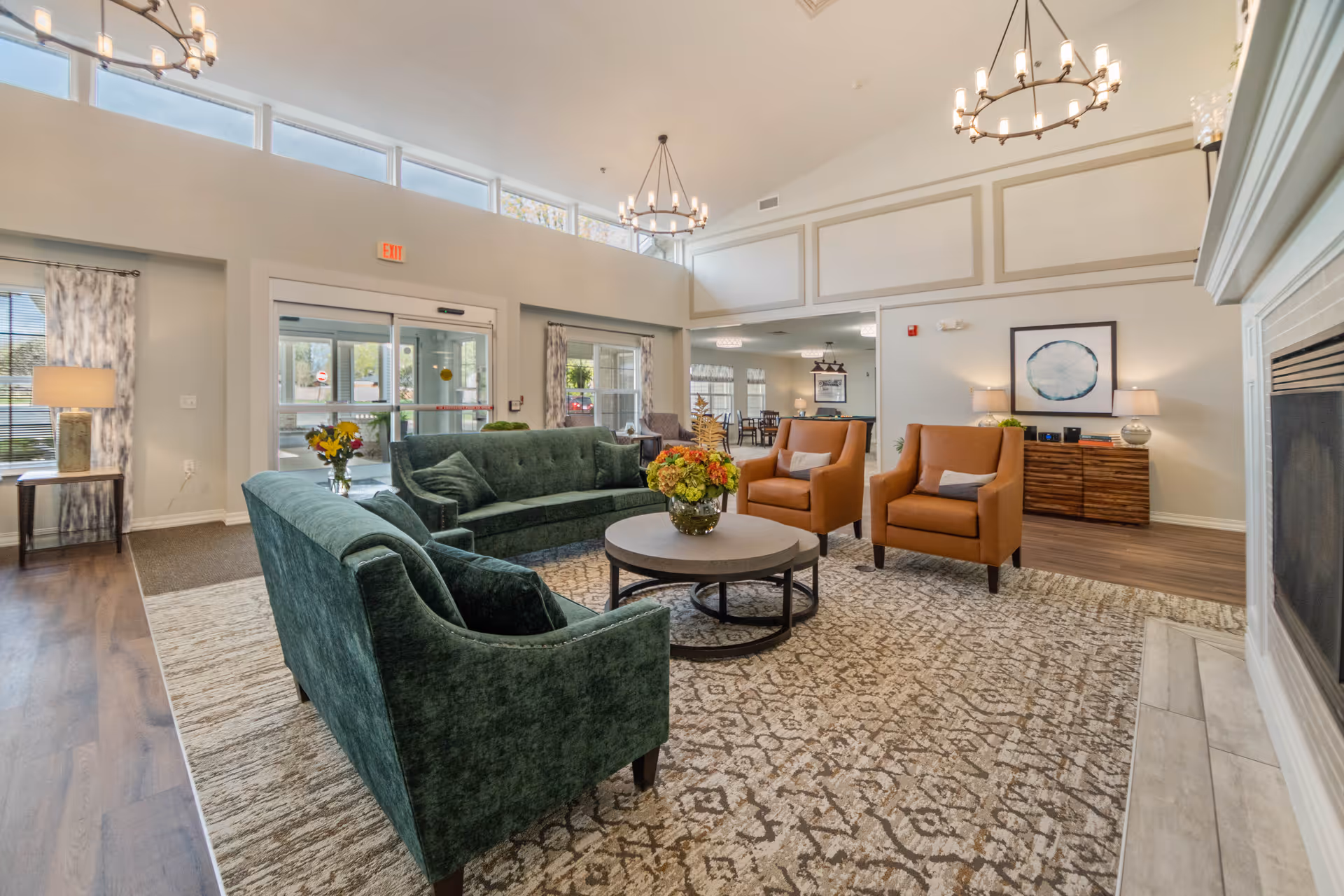 A bright and spacious living room area in a senior living facility with large windows near the ceiling allowing natural light. The room features a green velvet sofa, two matching green armchairs, two orange armchairs, a round coffee table with a flower arrangement, a patterned area rug, and a fireplace on the right. There are two chandeliers hanging from the ceiling, a wooden sideboard with lamps and decor, and a view into a dining area in the background.