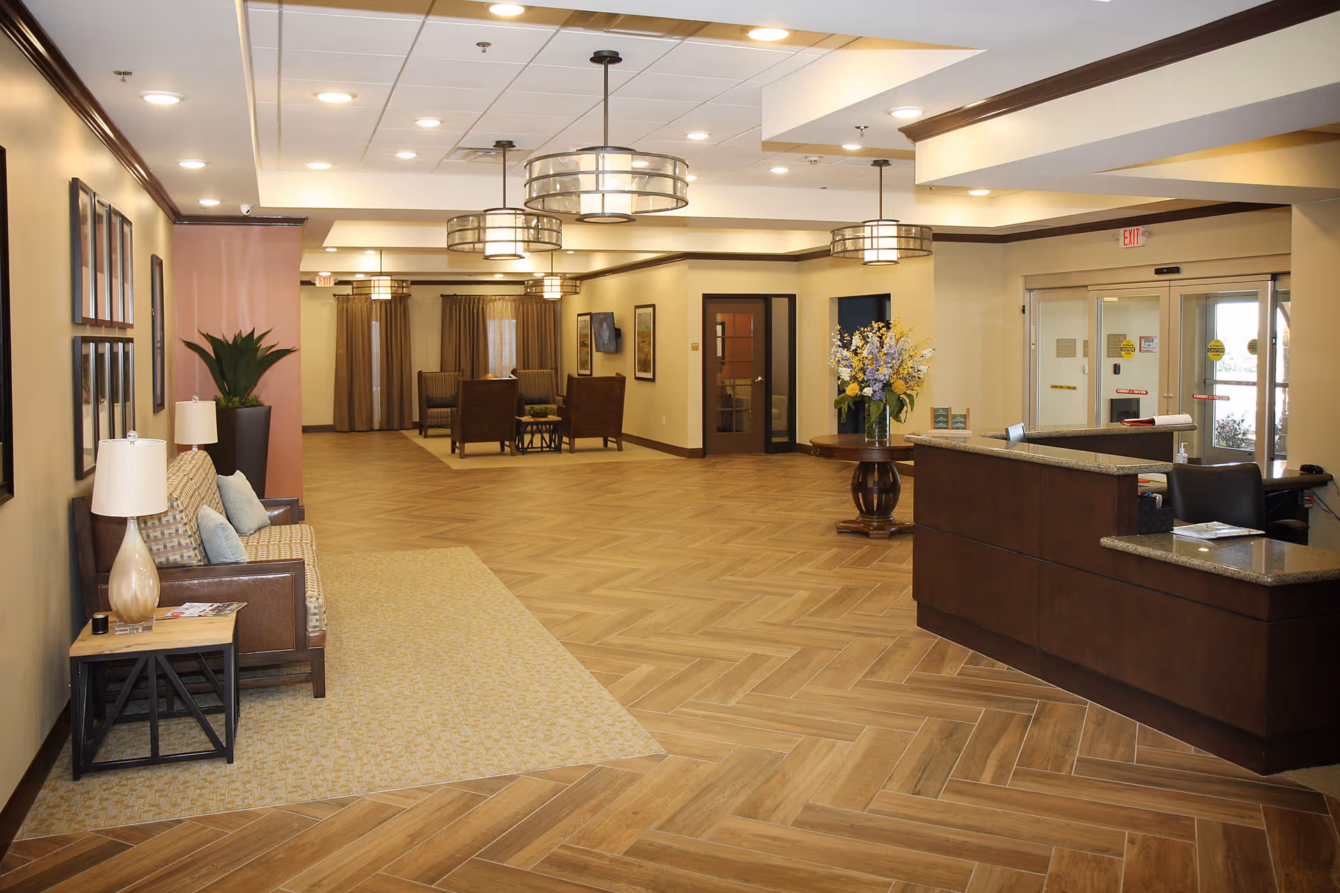 Spacious and well-lit assisted living facility lobby with a reception desk on the right, a seating area with a couch and lamps on the left, and additional seating with chairs and tables in the background. The floor has a wood pattern, and there are modern hanging light fixtures on the ceiling. A round table with a flower arrangement is near the entrance doors.