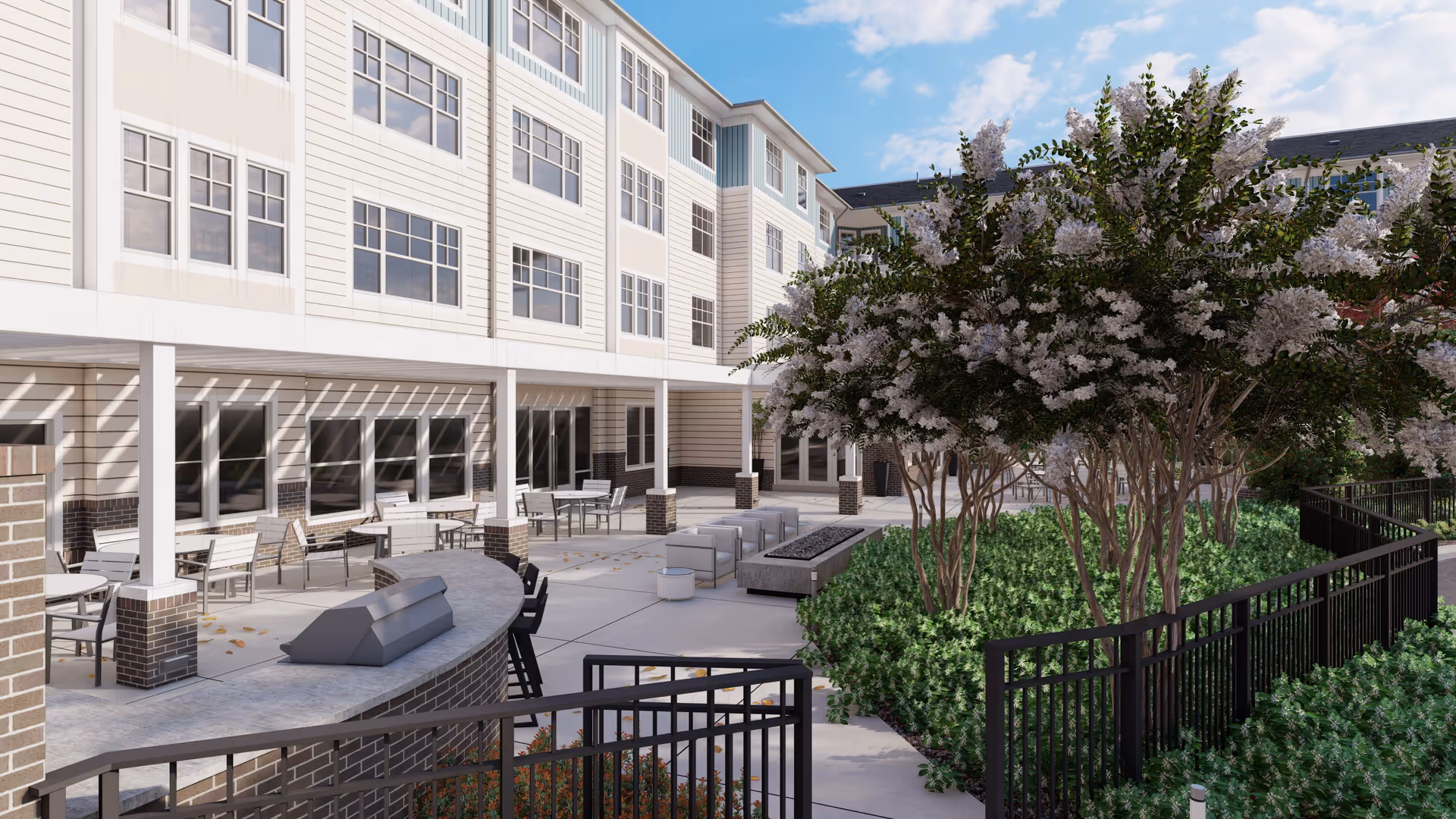 Outdoor patio area of a senior living facility with multiple seating arrangements including tables and chairs under a covered walkway, a curved counter with bar stools, and a fire pit surrounded by cushioned chairs. The area is landscaped with green bushes and trees with white flowers, enclosed by a black metal fence. The building exterior is light-colored with multiple windows and a blue sky with some clouds above.