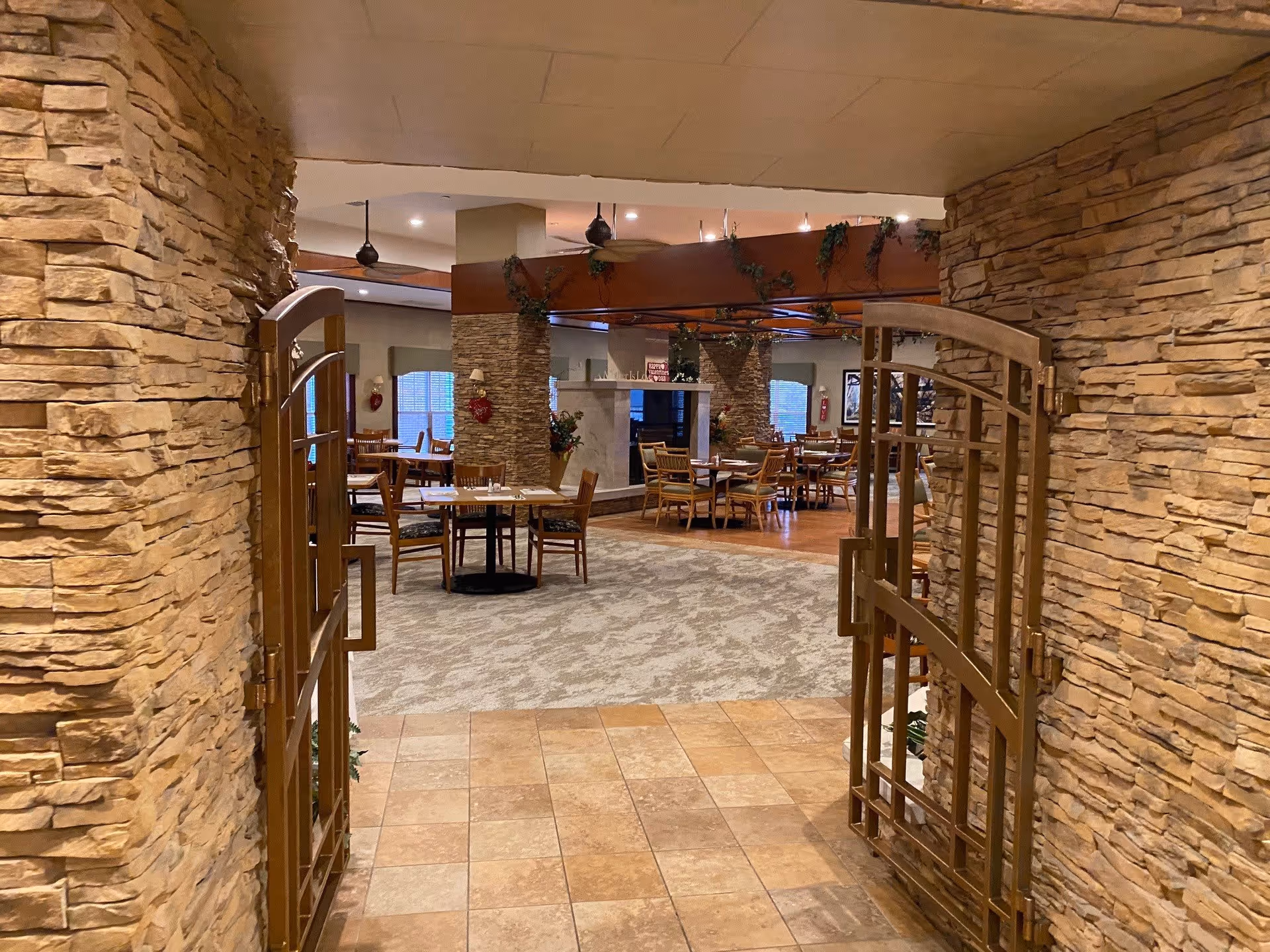 View through an open decorative metal gate into a dining area with multiple wooden tables and chairs. The room features stone walls, a tiled floor at the entrance, carpeted flooring in the dining area, ceiling fans, and a fireplace in the center. The space is warmly lit and decorated with some greenery.