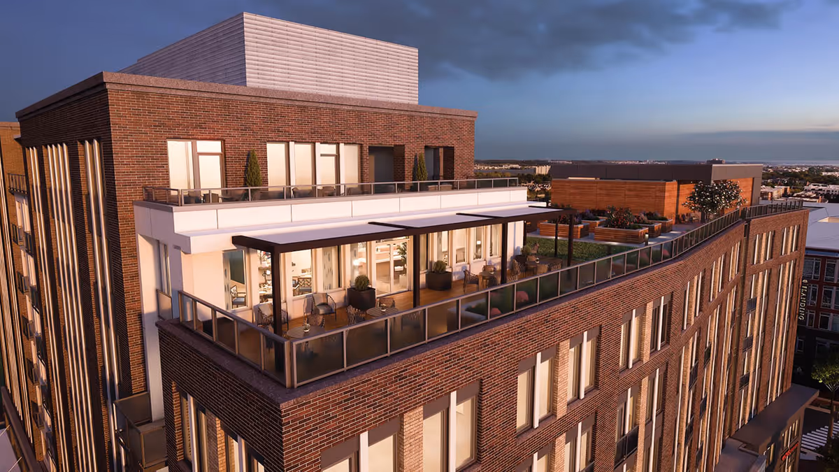 Exterior view of a modern multi-story brick building at dusk with a spacious rooftop terrace featuring outdoor seating, plants, and a pergola.